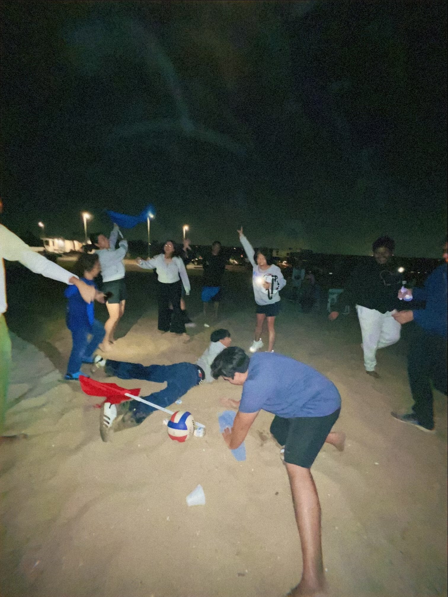 People playing volleyball on a sandy beach at night, with some individuals holding items and others involved in the game, under streetlights.