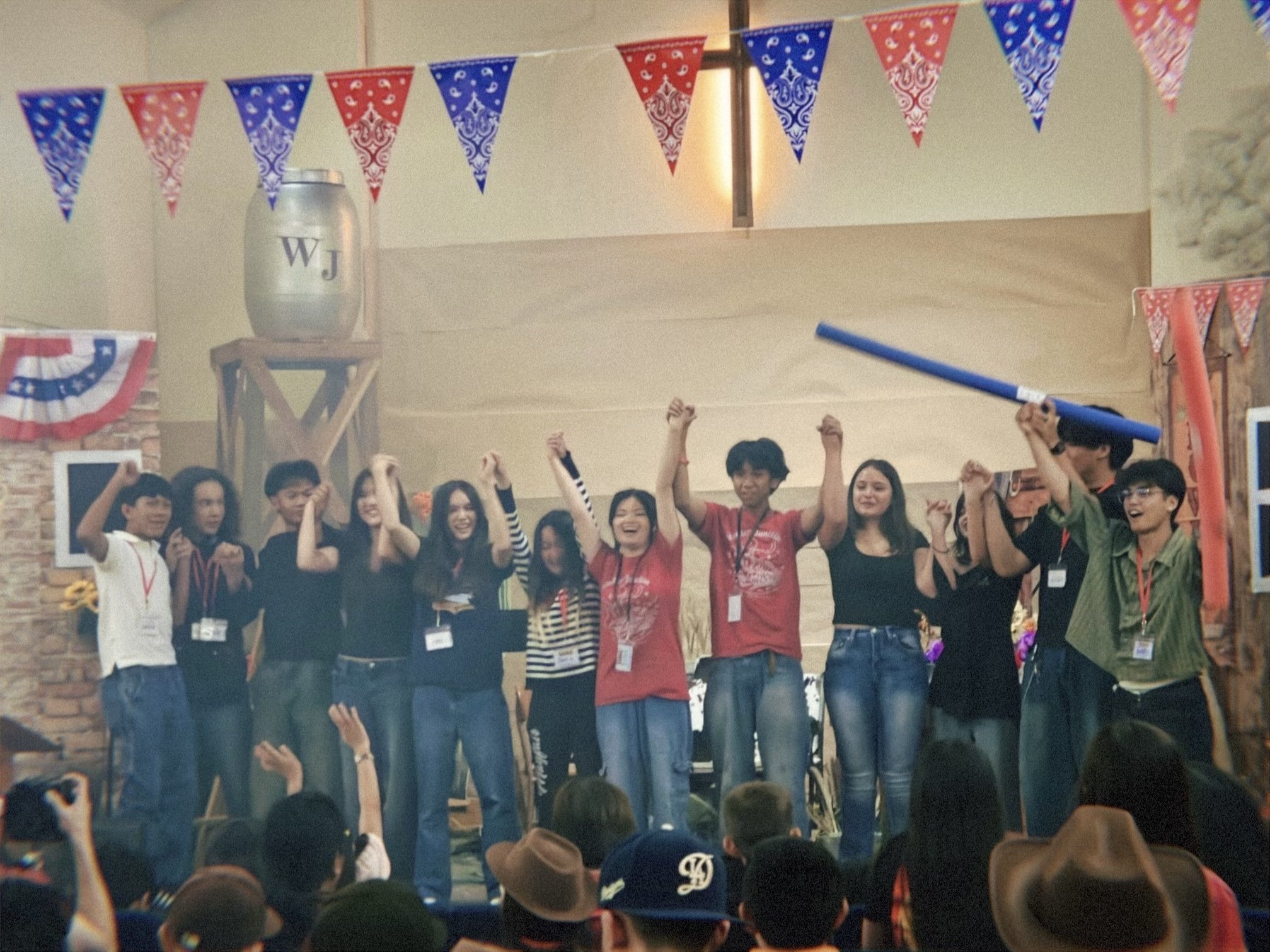 Group of people celebrating indoors, raising their hands, with red, white, and blue patriotic bunting decorations hanging from the ceiling.