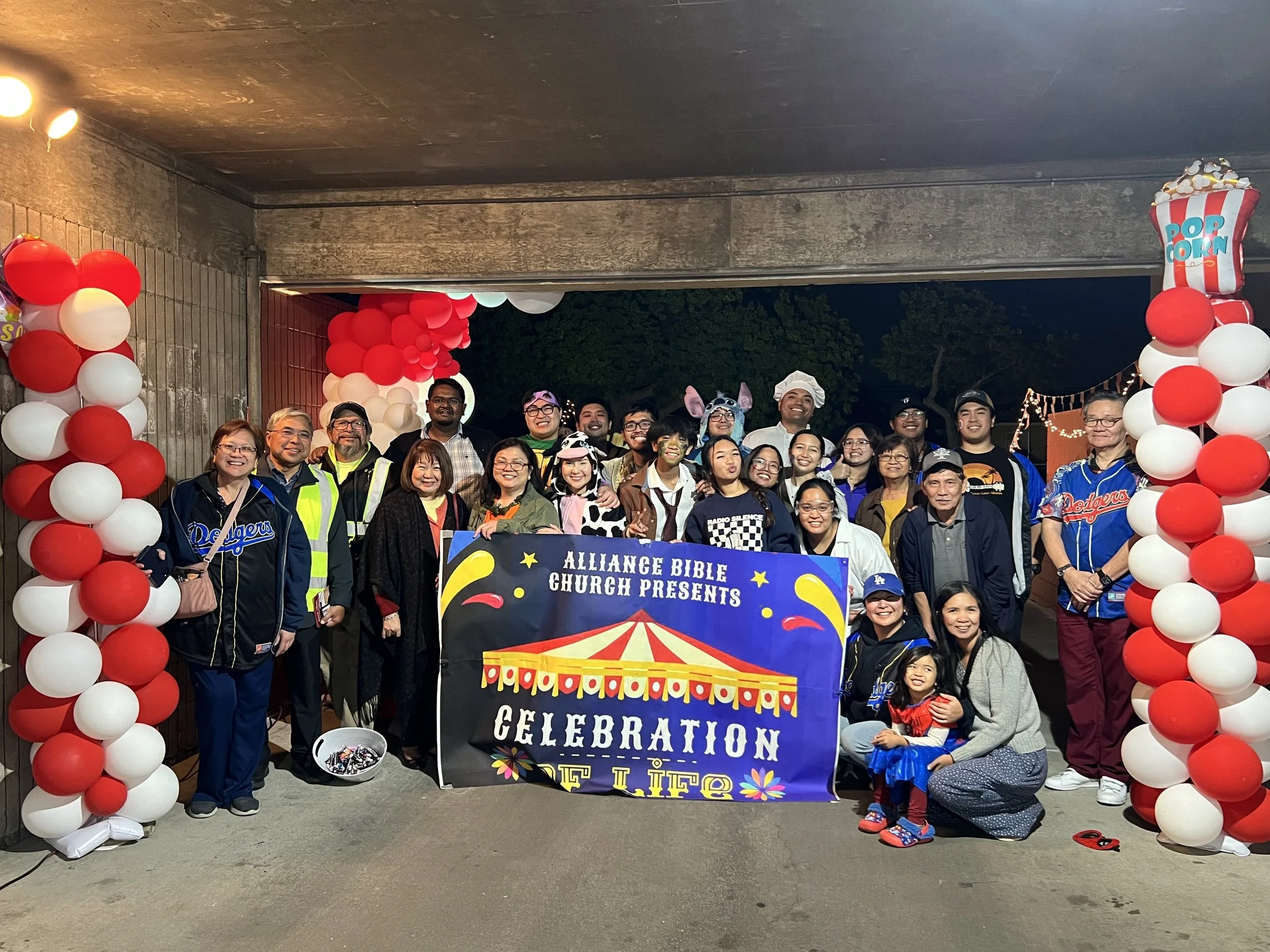 Group of people at an outdoor celebration under a decorated archway of red and white balloons, holding a colorful banner that reads 'Alliance Bible Church Presents, Celebration of Life'.