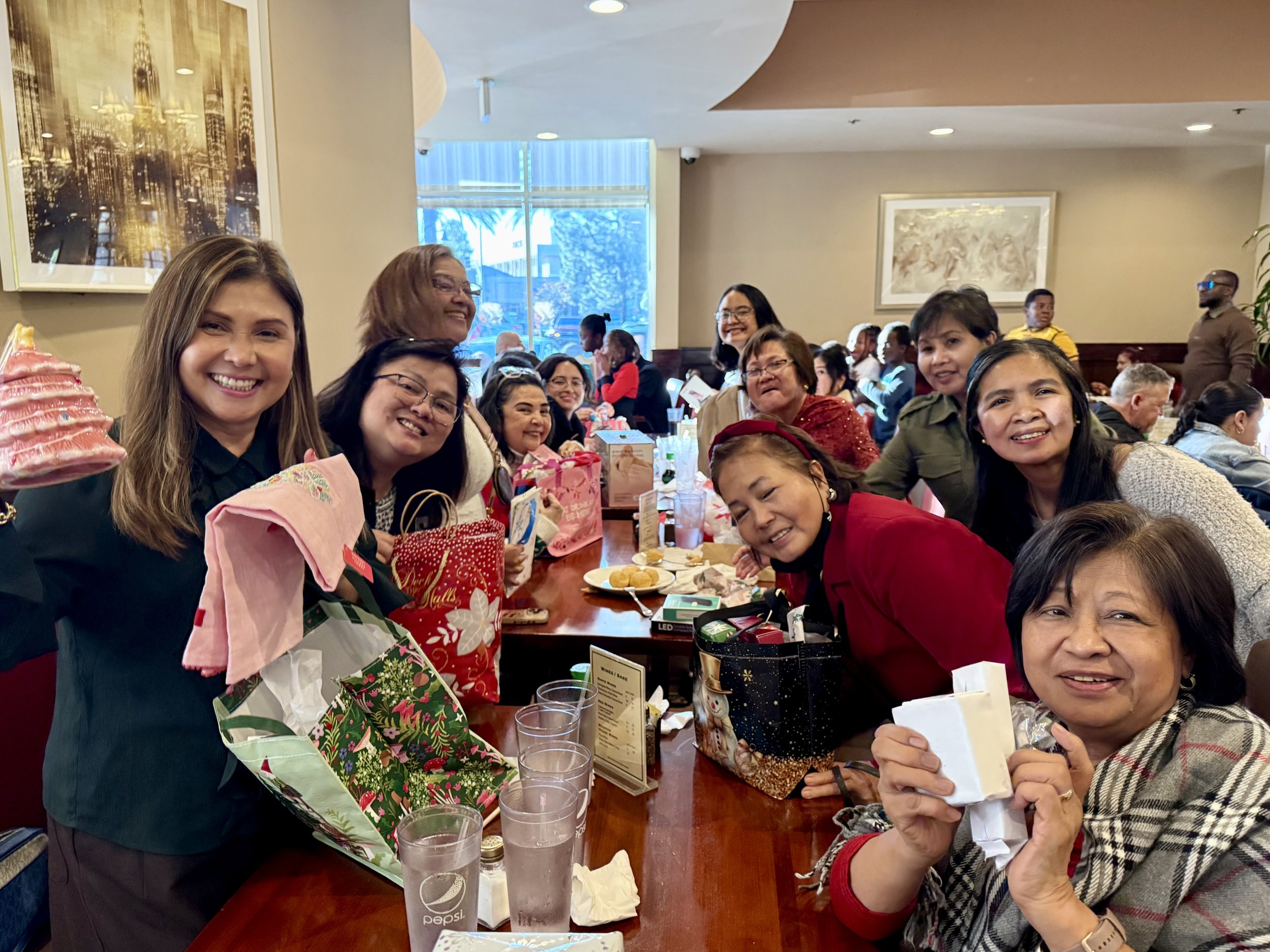 A group of smiling women and girls gathered around a table at a celebration, with gift bags and food on the table, in a restaurant or banquet hall.