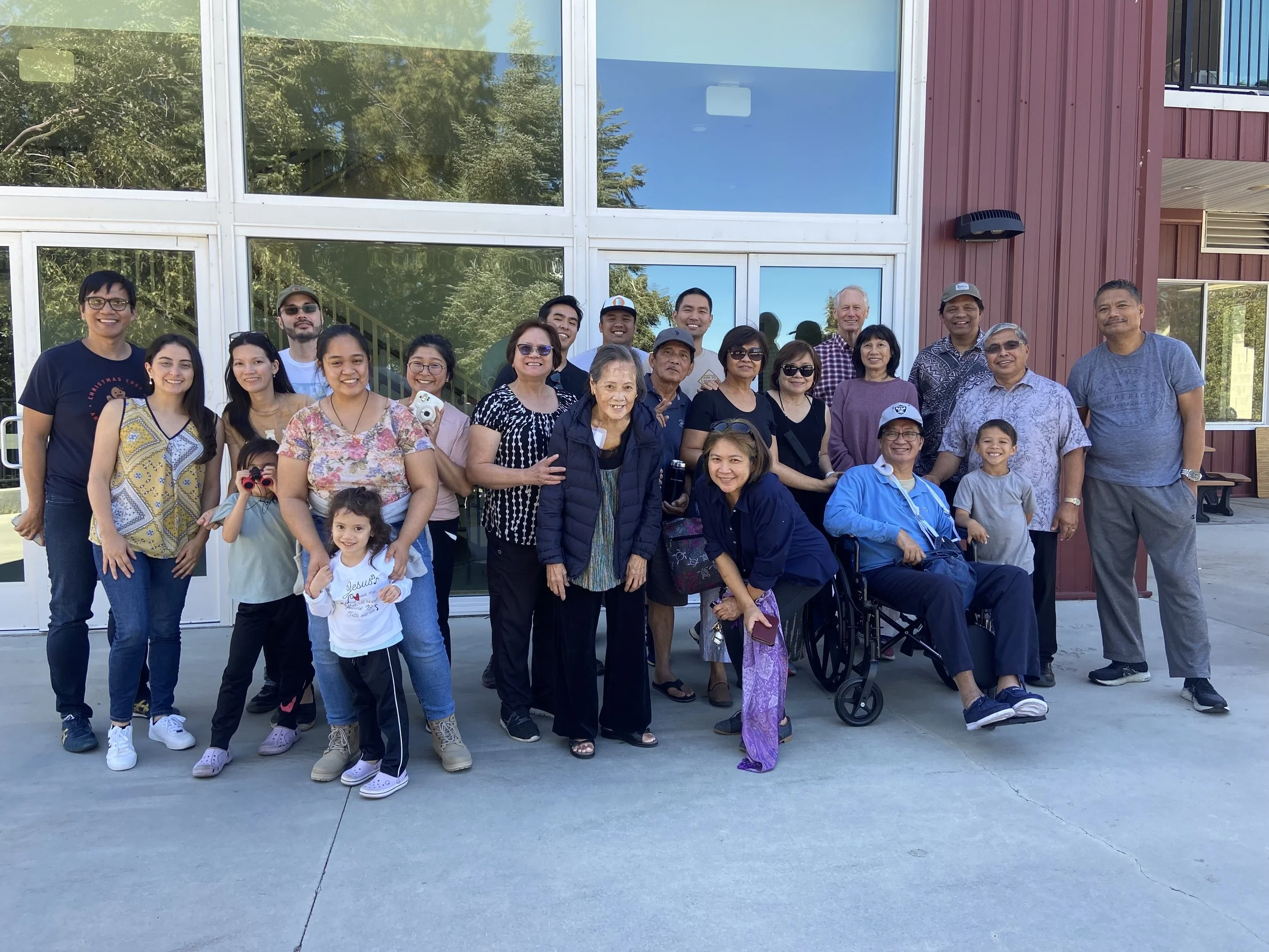 A group of diverse people, including children and elderly, posing for a photo outside a modern building with large glass windows. They are standing on a concrete surface with trees reflected in the windows.