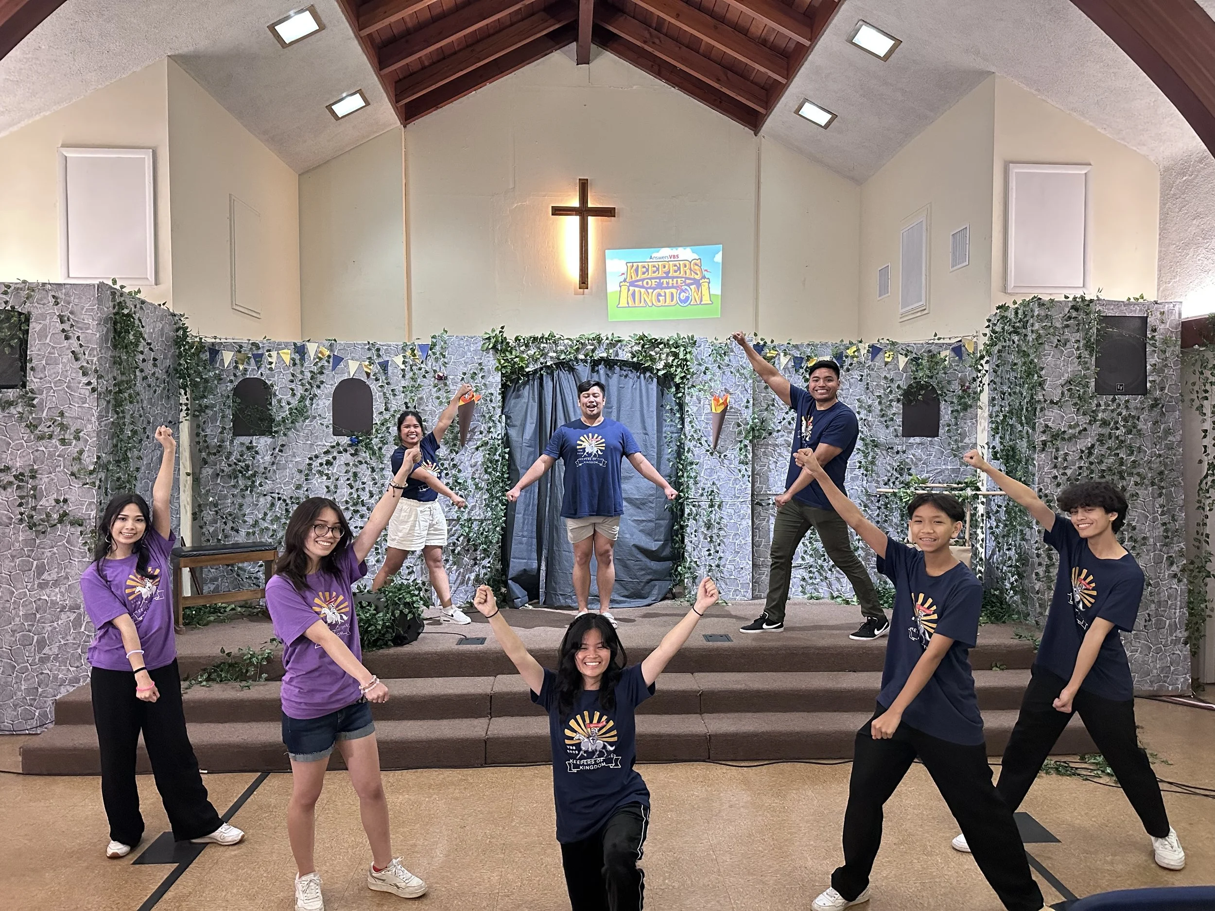 Group of smiling young people posing on stage with a Christian cross and a projector screen that reads "Keepers of the Kingdom". They are wearing blue and purple T-shirts. The stage has a stone wall backdrop decorated with greenery.