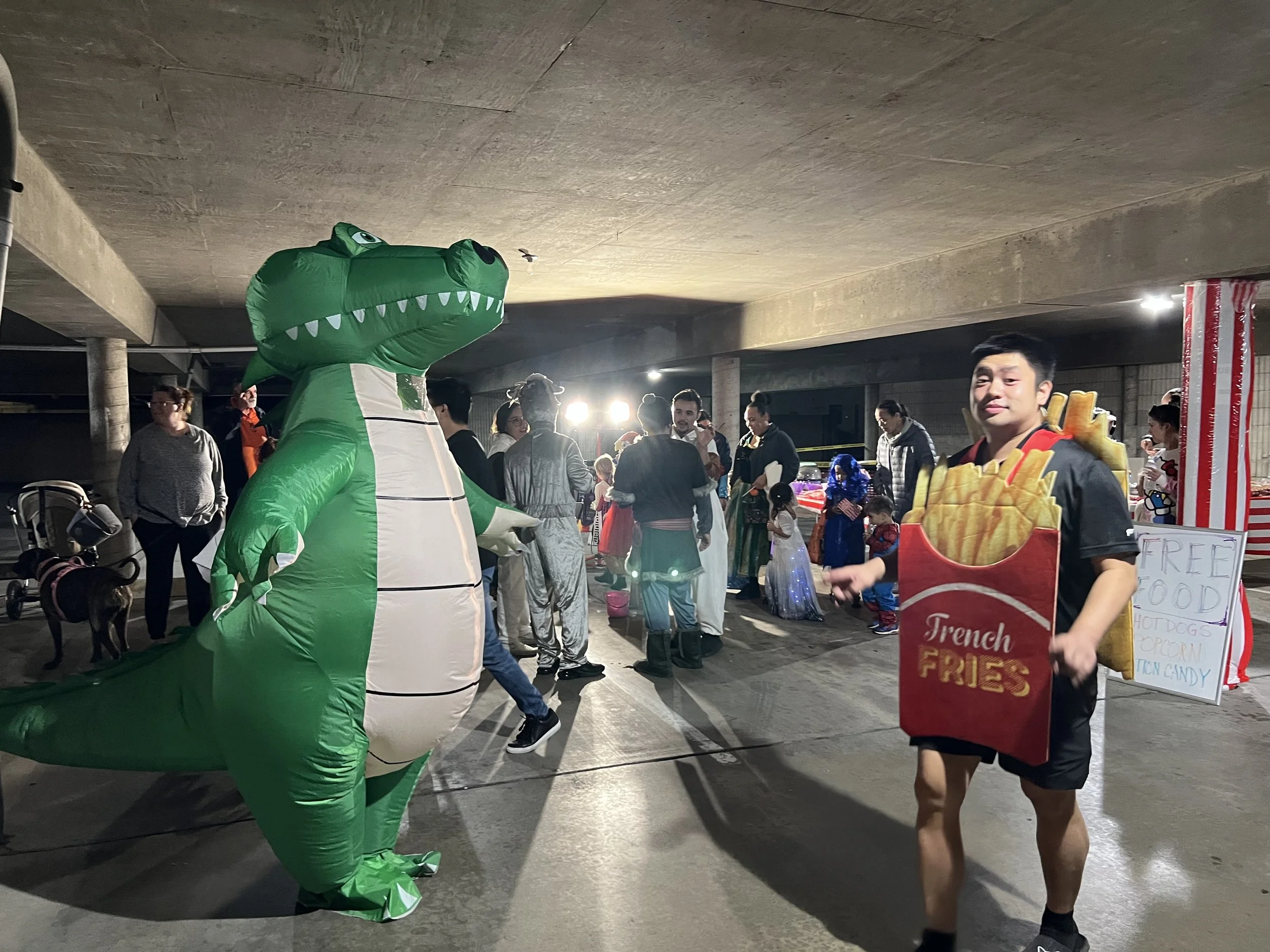 People at an indoor event with a person in an inflatable green dinosaur costume. A man is dressed as French fries holding a sign offering free food. The background shows other attendees and vendor stalls.