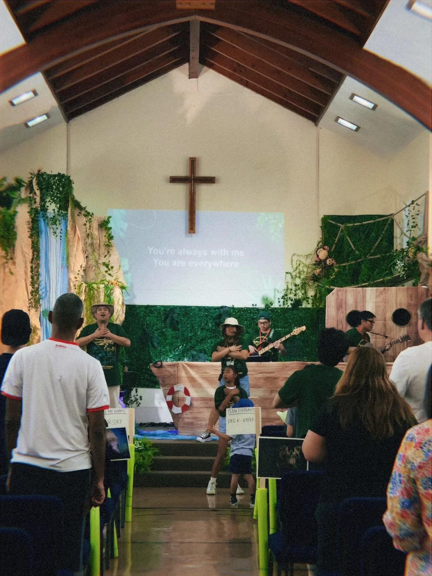 A church stage with a wooden cross on the wall, decorated with plants and a backdrop of greenery and blue drapes. Several children are performing or participating on the stage, with some holding microphones or playing instruments. Audience members are watching the event.