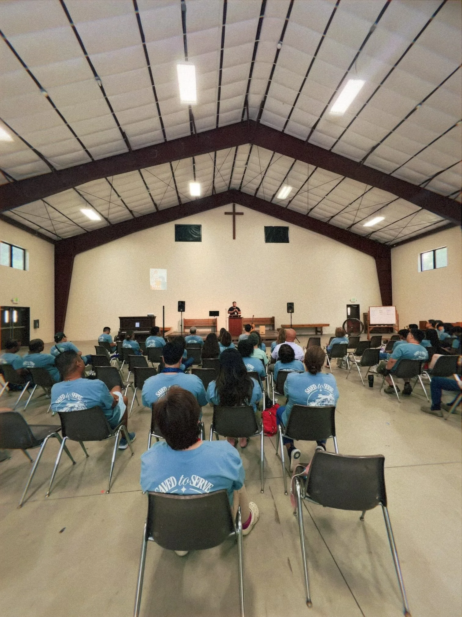 A church hall filled with people sitting on chairs facing a stage, with a person standing and speaking at the podium. The hall has a high ceiling with booth lighting and a large cross on the wall behind the speaker.