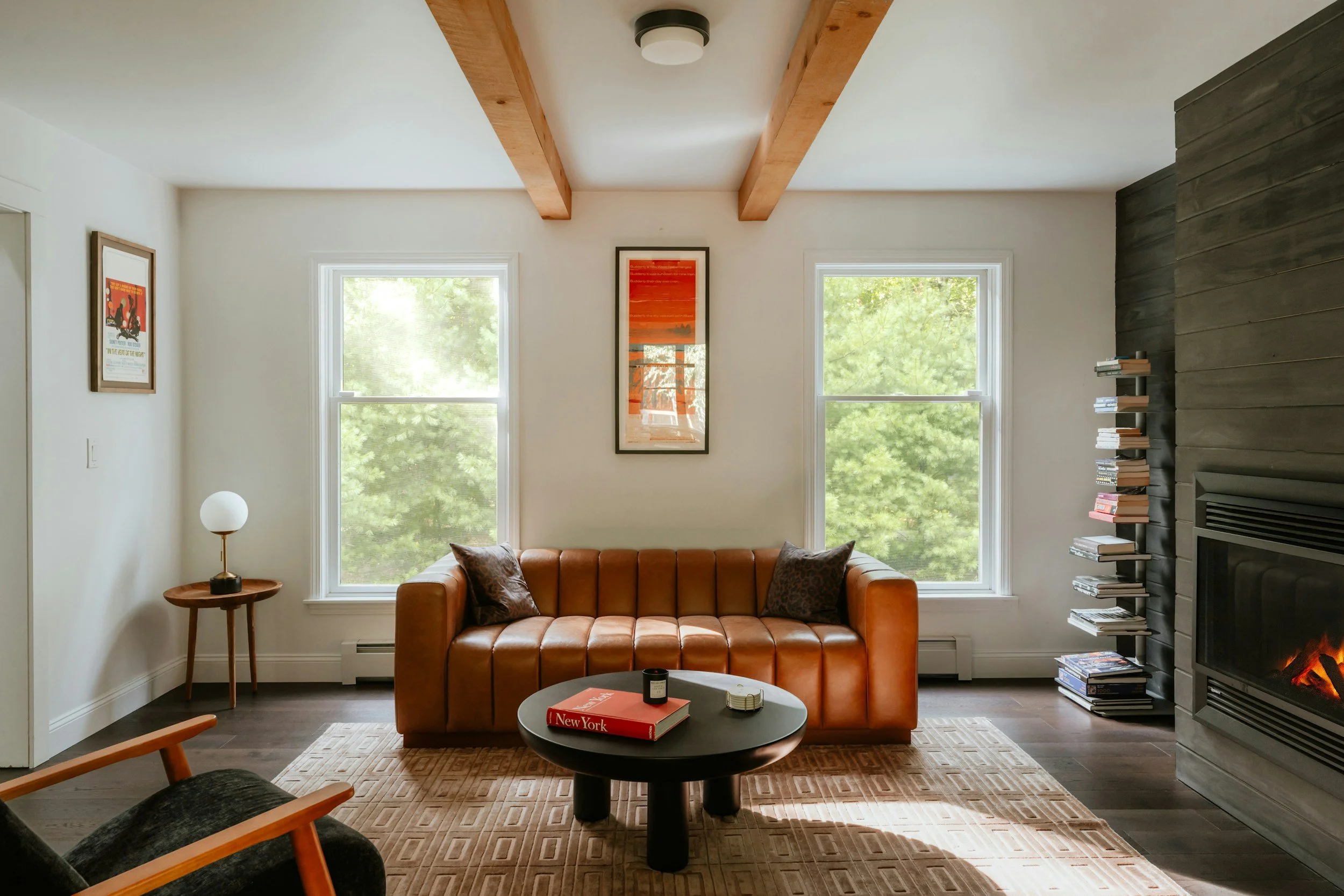Living room with tan leather sofa, two windows with green trees outside, a black round coffee table with a book and candle, a fireplace with a stack of books beside it, beige rug, wooden beam ceiling, and framed art on the wall.
