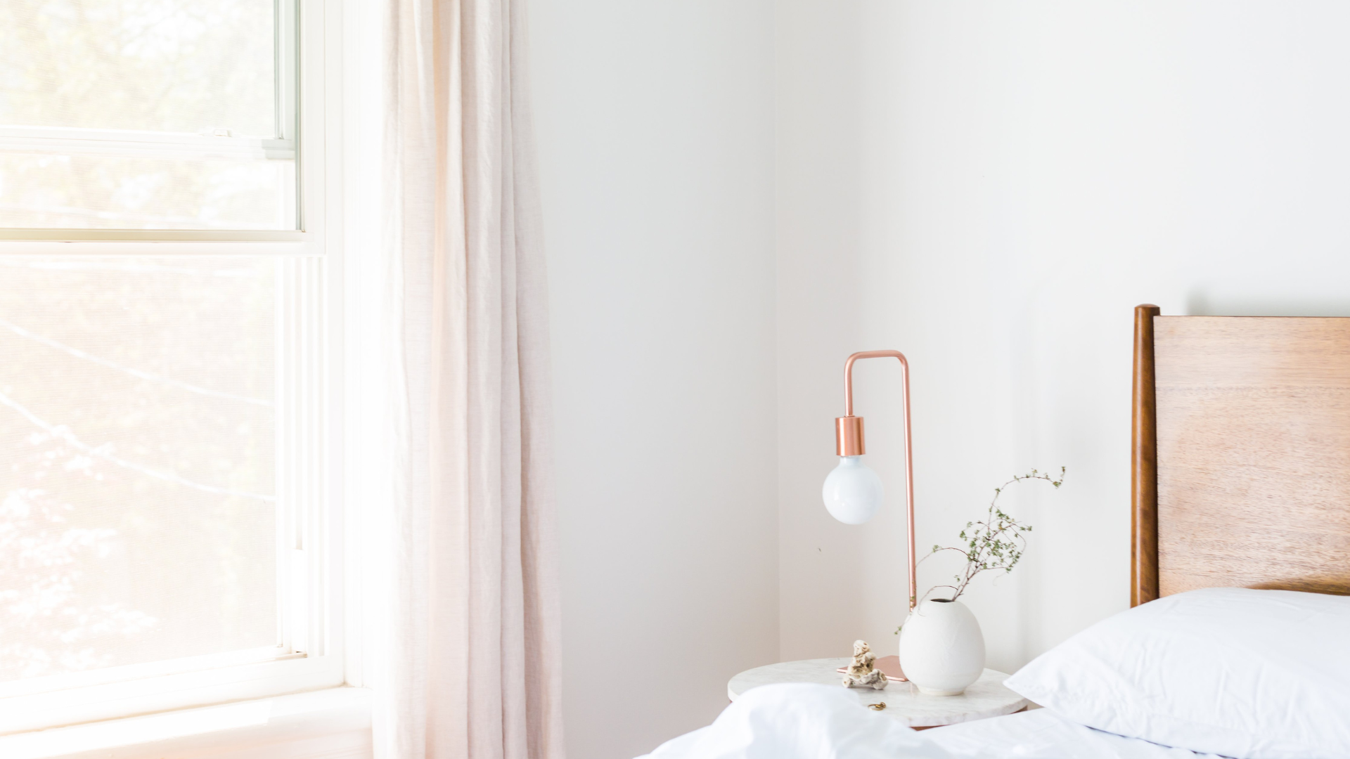 A bedroom corner with a wooden bed frame, white bedding, a round bedside table, a white vase with a sprig of greenery, and a pink copper wall lamp near a window with light-colored curtains.