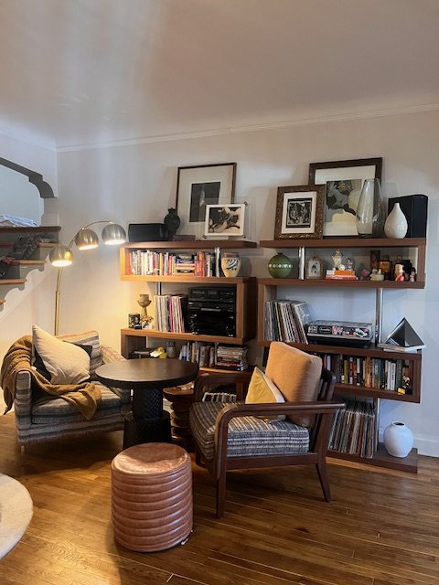Living room with a patterned sofa, wooden armchair with yellow cushion, round side table, tufted ottoman, and wooden shelves with books, framed pictures, and decorative objects.