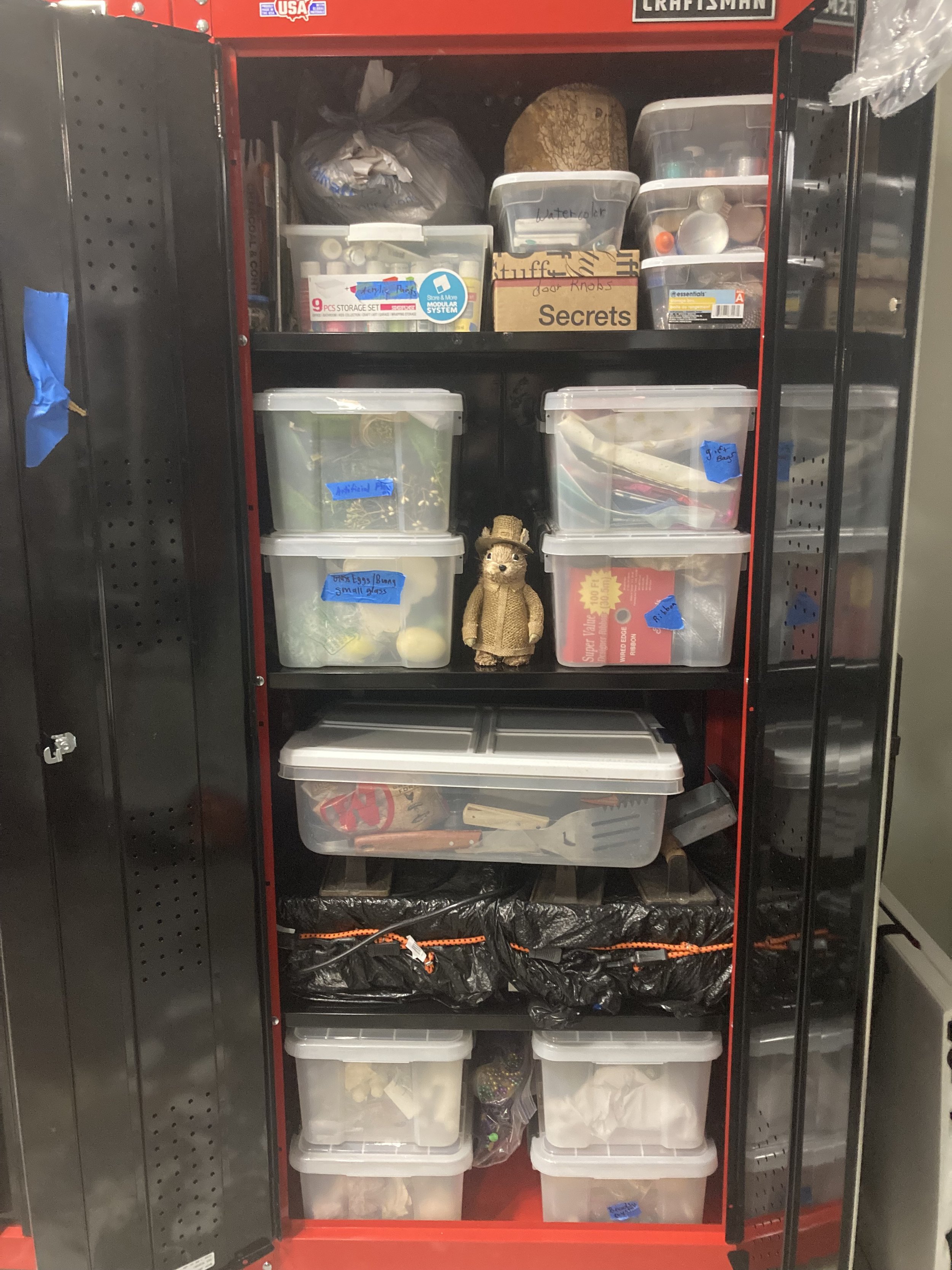 A red metal storage cabinet filled with plastic bins, jars, and containers, containing various tools, craft supplies, and miscellaneous items. There are labels and a stuffed bear toy on the shelves.