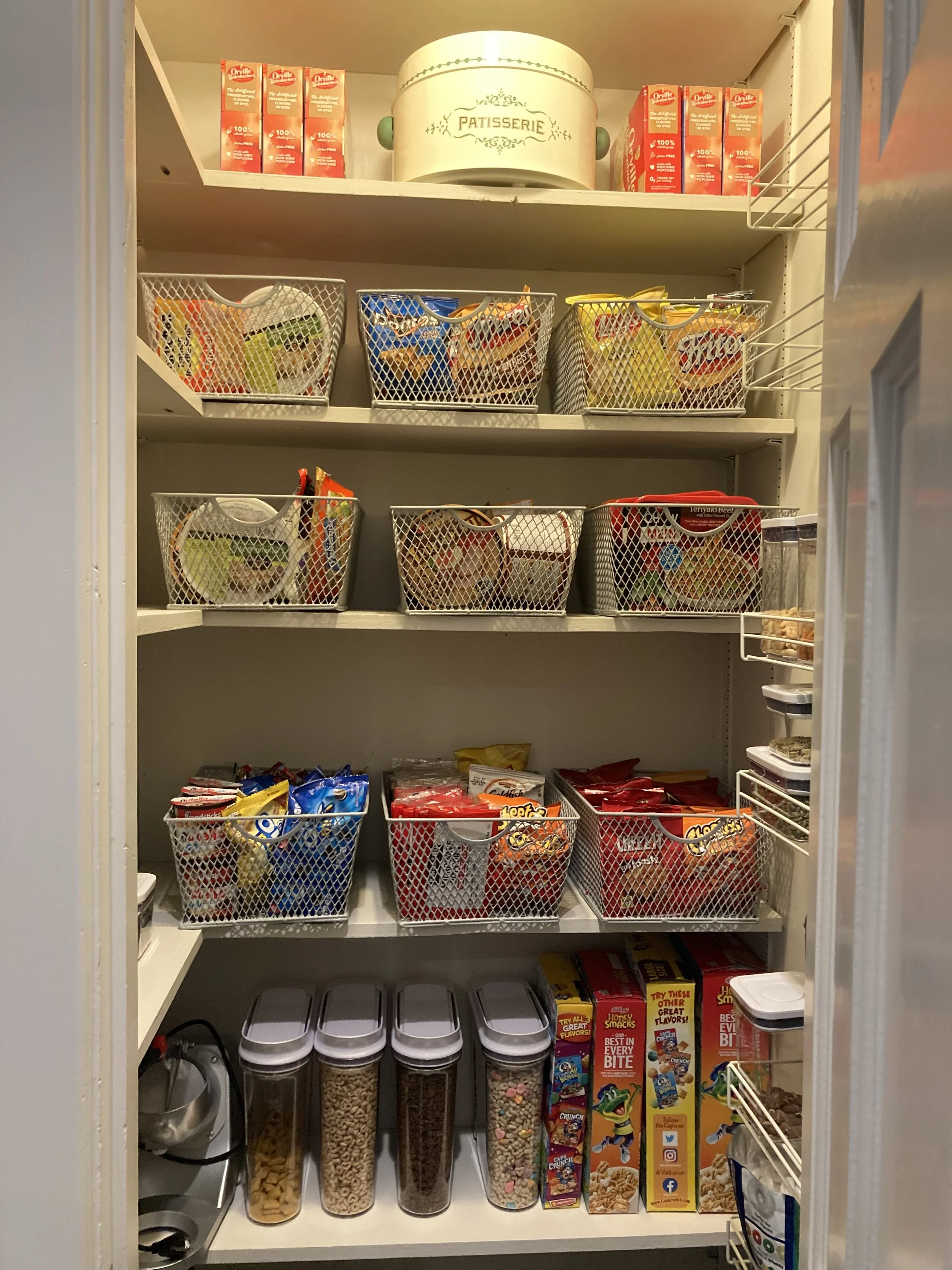 Pantry shelf filled with snack foods including chips, cereals, and crackers in various containers and boxes, with a decorative pastry tin labeled "Patisserie" at the top.