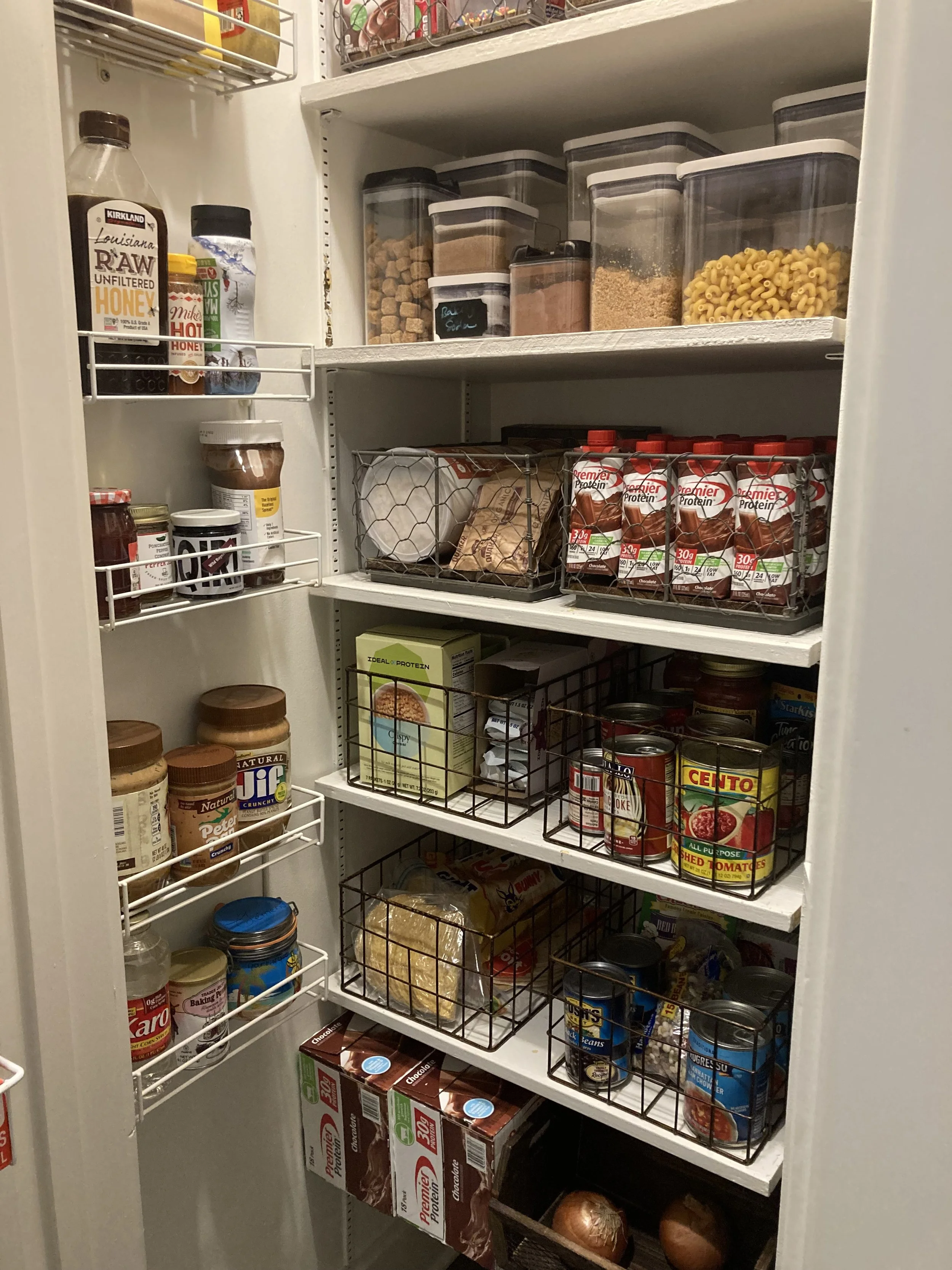 Inside a well-organized pantry with shelves containing jars, cans, boxes, and sealed food containers, including pasta, canned goods, protein shakes, spices, and snacks.