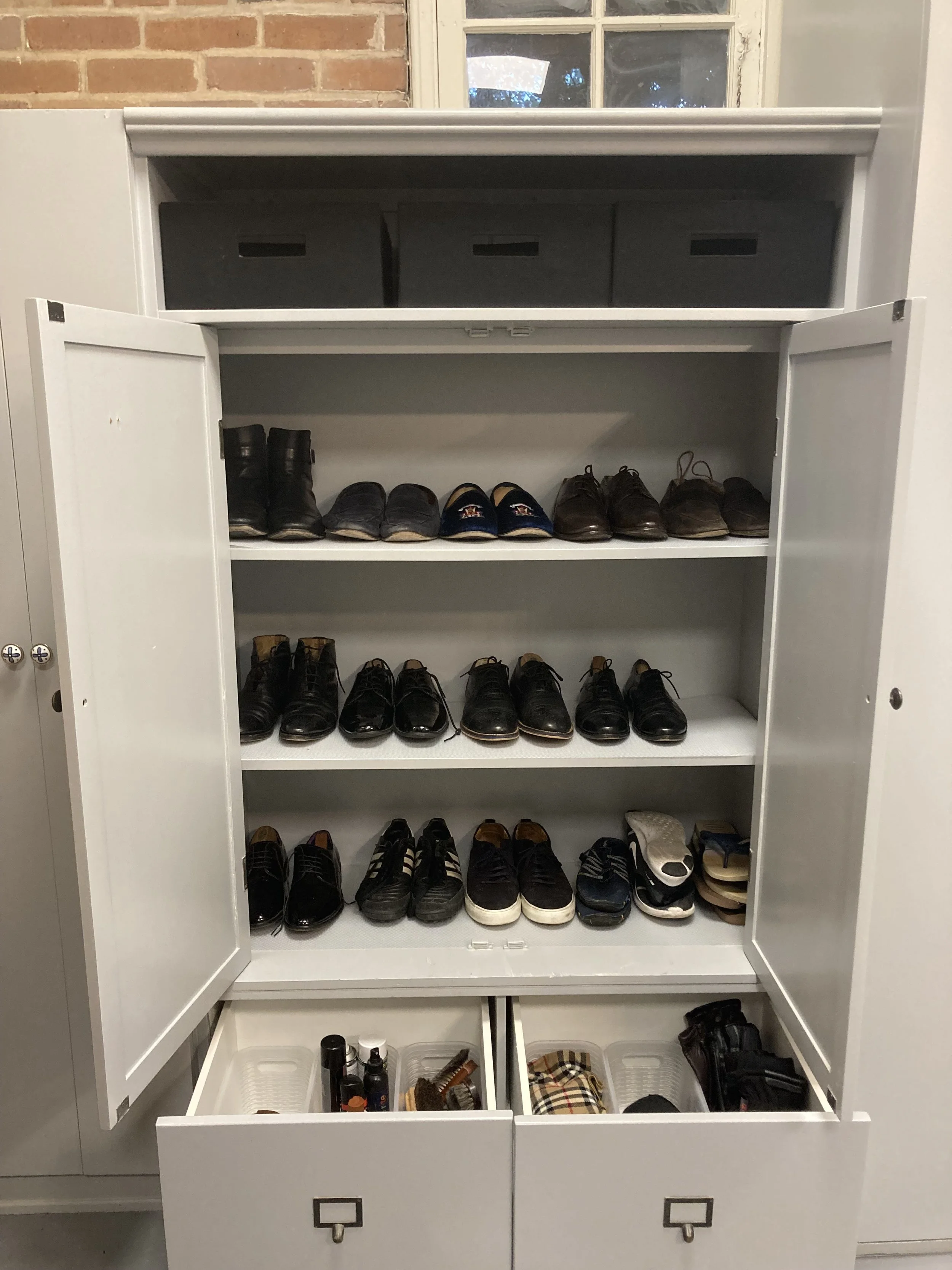 A cabinet with three shelves filled with men's shoes, organized by pairs, and the top shelf has black storage boxes. The cabinet is open, revealing shoes in various styles, with some drawers at the bottom containing grooming products and other accessories.