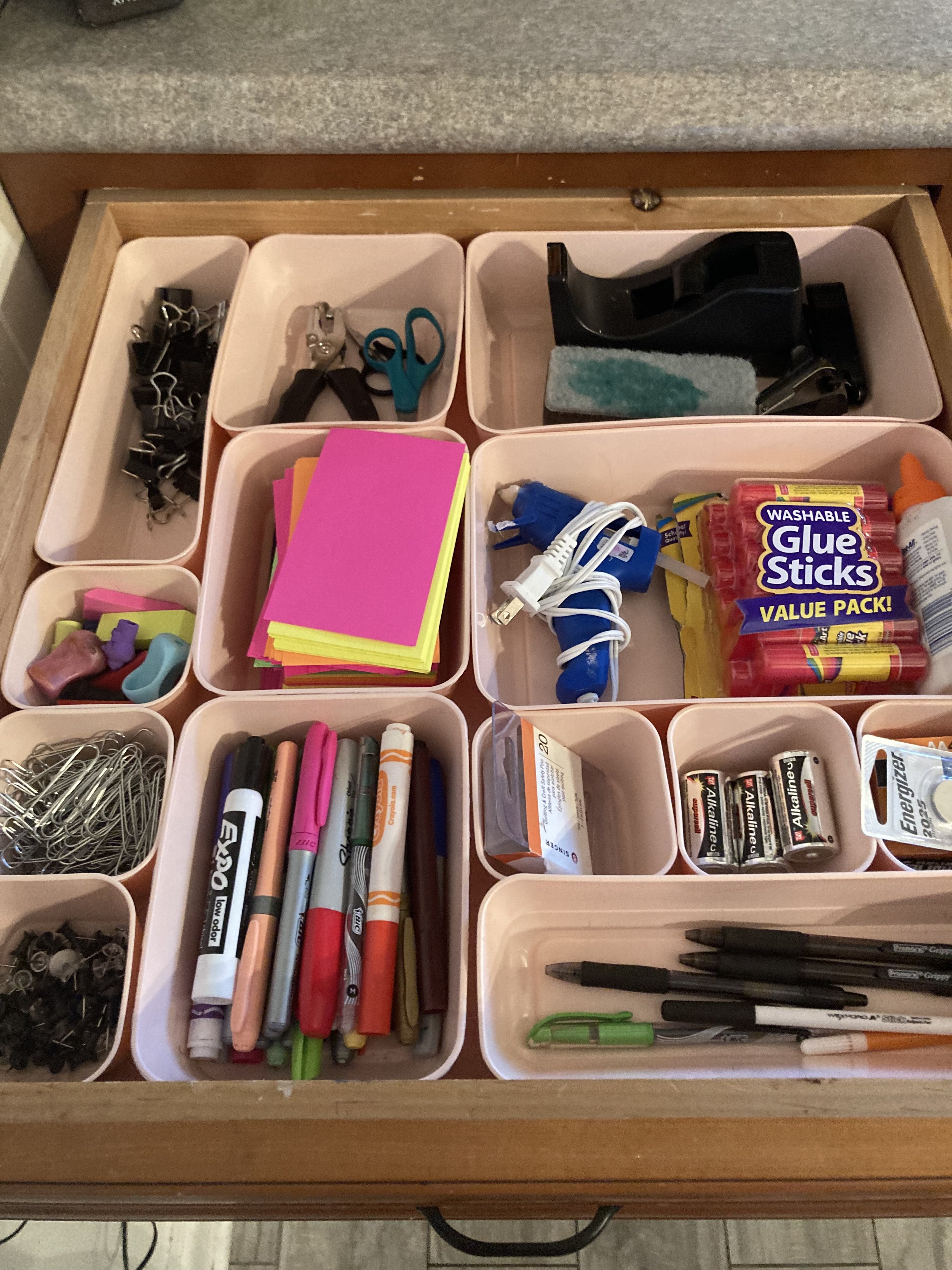 Organizer drawer containing office supplies such as paper clips, scissors, stapler, glue sticks, batteries, pens, markers, push pins, markers, small containers, and a highlighter.