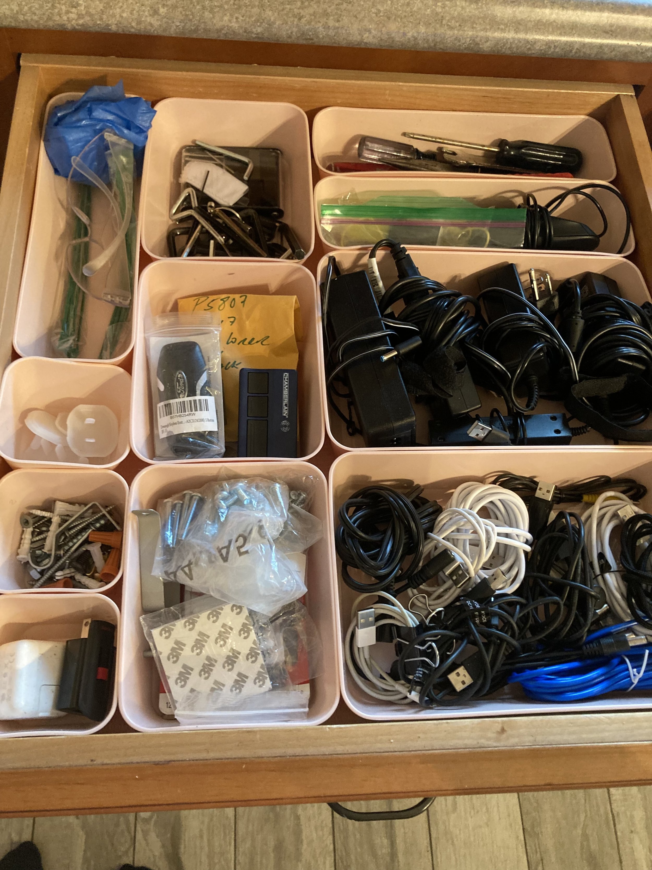 A wooden drawer with multiple small plastic compartments filled with various electronic cables, chargers, screws, and small hardware accessories.