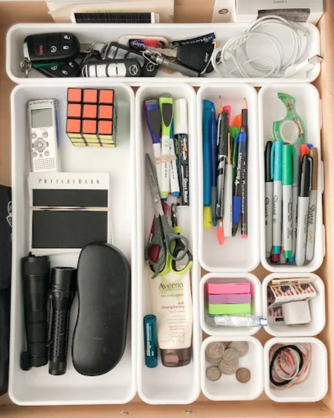 Drawer organizer with miscellaneous items including remote controls, a Rubik's cube, pens, scissors, a flashlight, a tube of skin cream, sticky notes, coins, and assorted small objects.