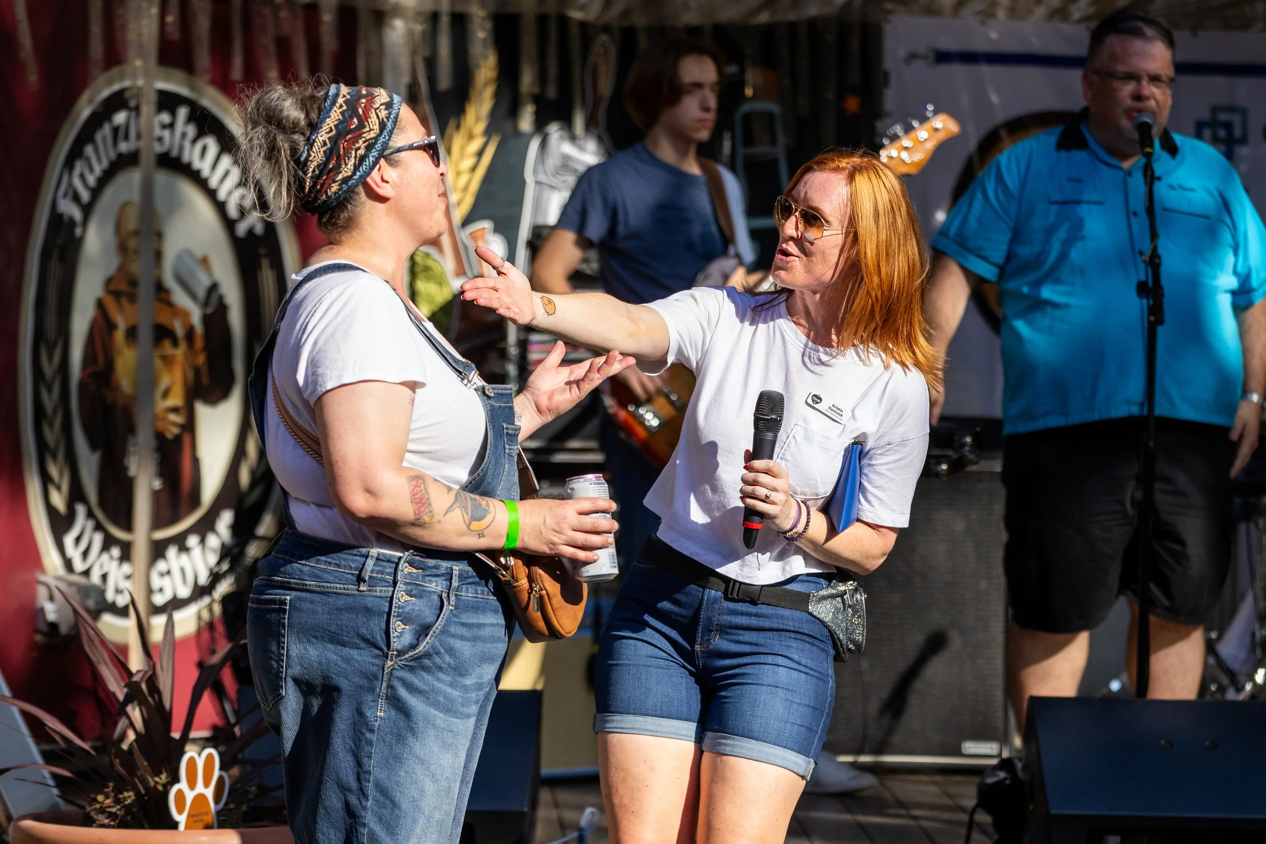 Two women are engaged in a conversation on stage at an outdoor event, one holding a microphone and wearing sunglasses, while the other has tattoos and is only in profile. There are band members in the background, including a guitarist and a man singing into a microphone, with stage equipment and a logo visible.