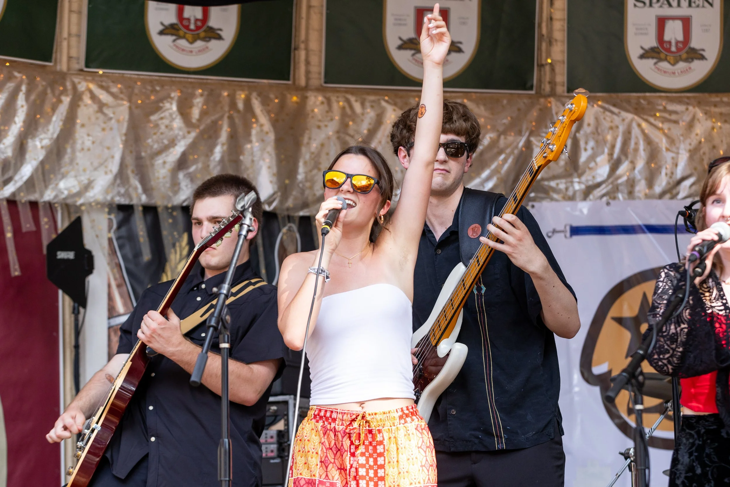 A female singer with dark hair, sunglasses, and a white top singing into a microphone, raising one arm, accompanied by male guitar players at a live outdoor concert.