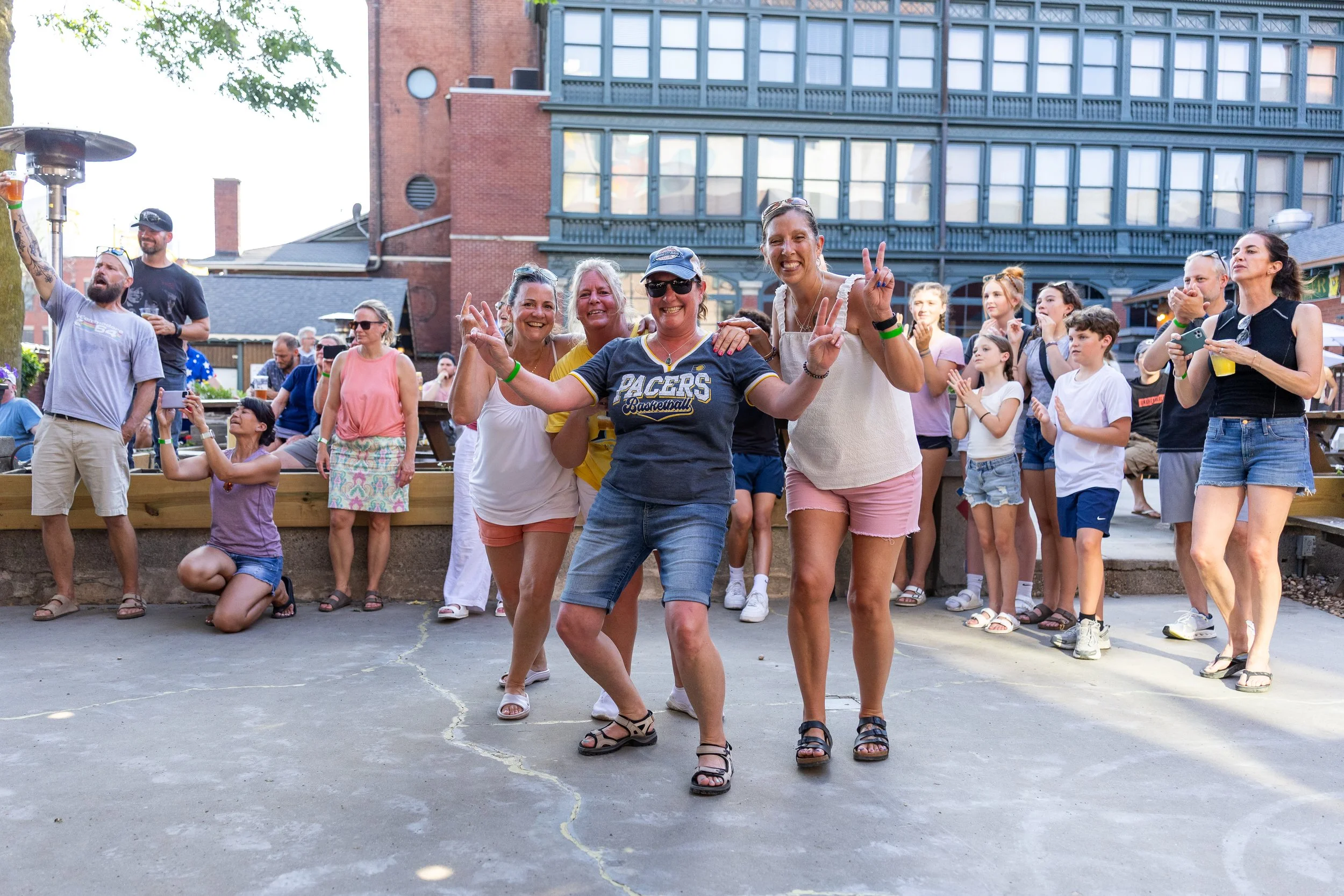 Group of people enjoying an outdoor event, smiling and making peace signs, with a brick building in the background.