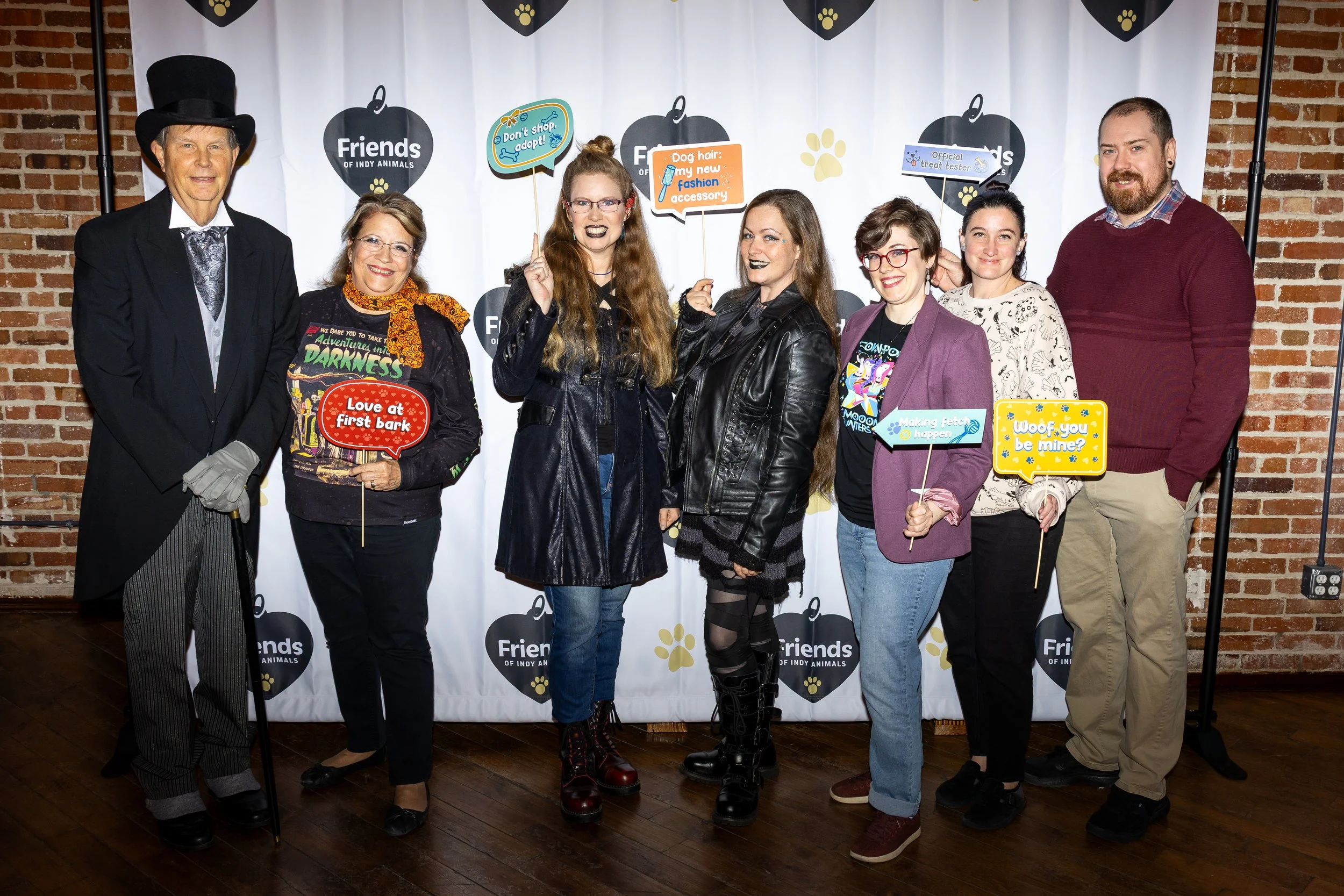 Group of seven people standing in front of a backdrop with the 'Friends of Indy Animals' logo. They are dressed in quirky outfits, holding fun signs related to dogs, pets, and adoption, with a brick wall to the left.
