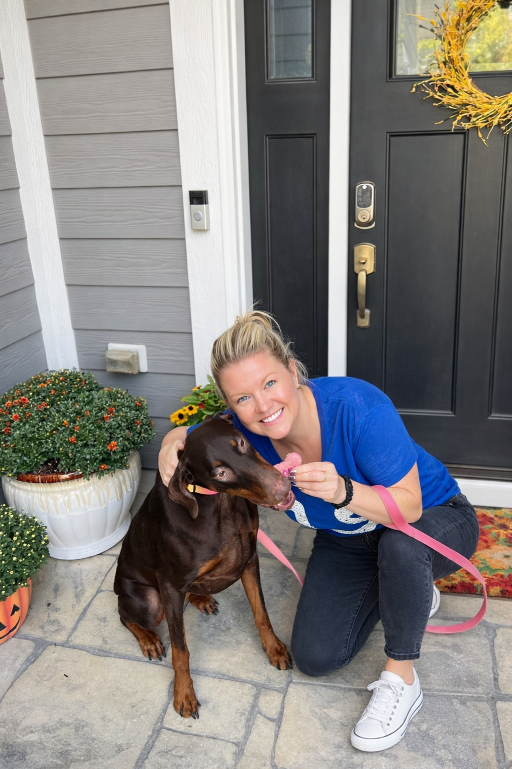 A woman in a blue shirt kneeling with a dog on a leash outside a house, holding a pink treat close to the dog's mouth, with a gray house wall, a black door, and potted flowers nearby.