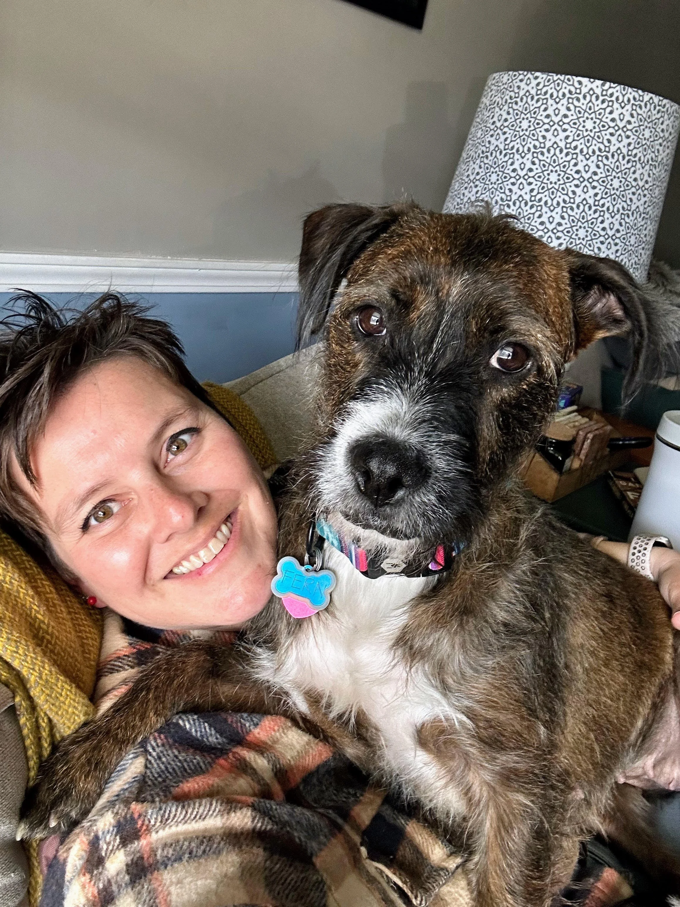 A woman smiling with a brindle-coated dog lying on her lap, wearing a colorful collar with tags, in a cozy indoor setting.