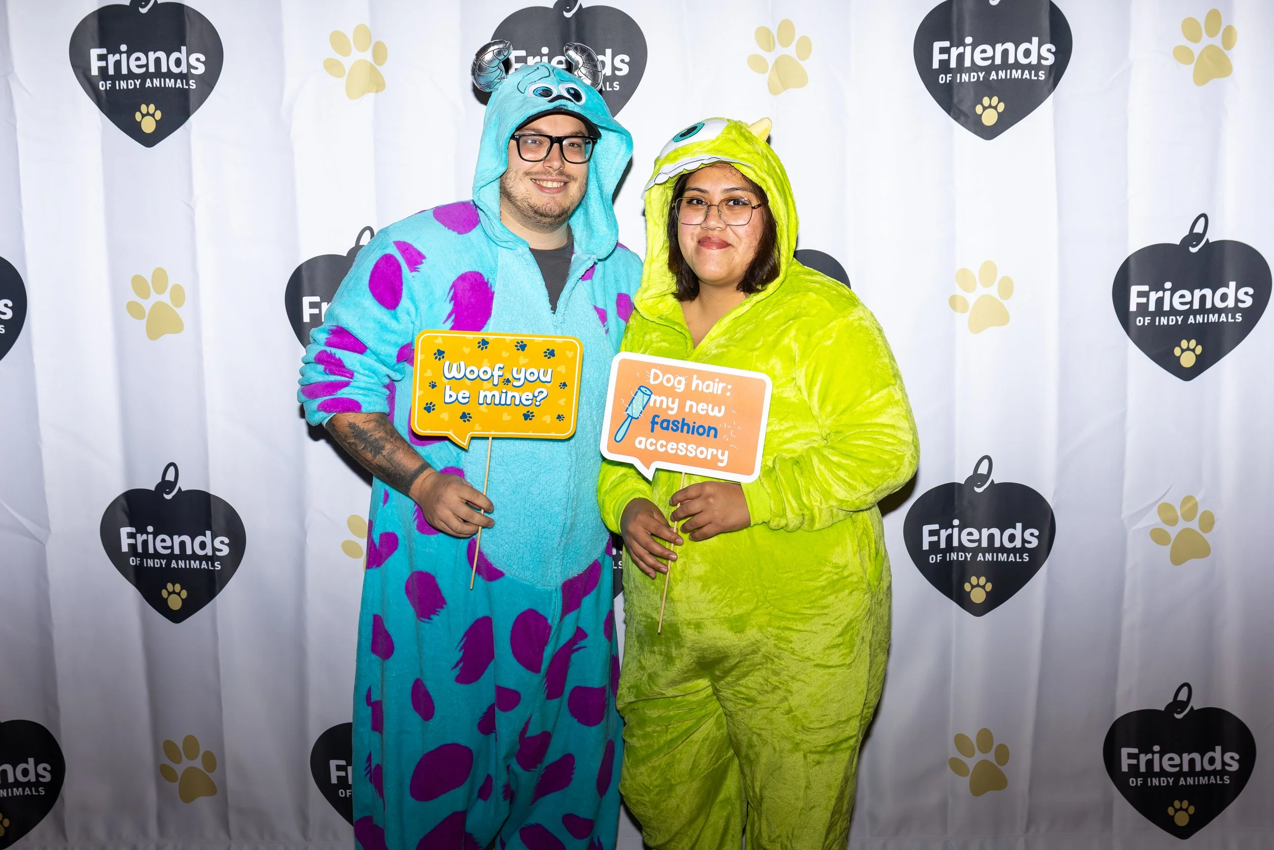 Two people dressed in colorful monster costumes standing in front of a backdrop with the Friends of Indy Animals logo. They are holding signs that say "Woof you be mine?" and "Dog hair: my new fashion accessory".