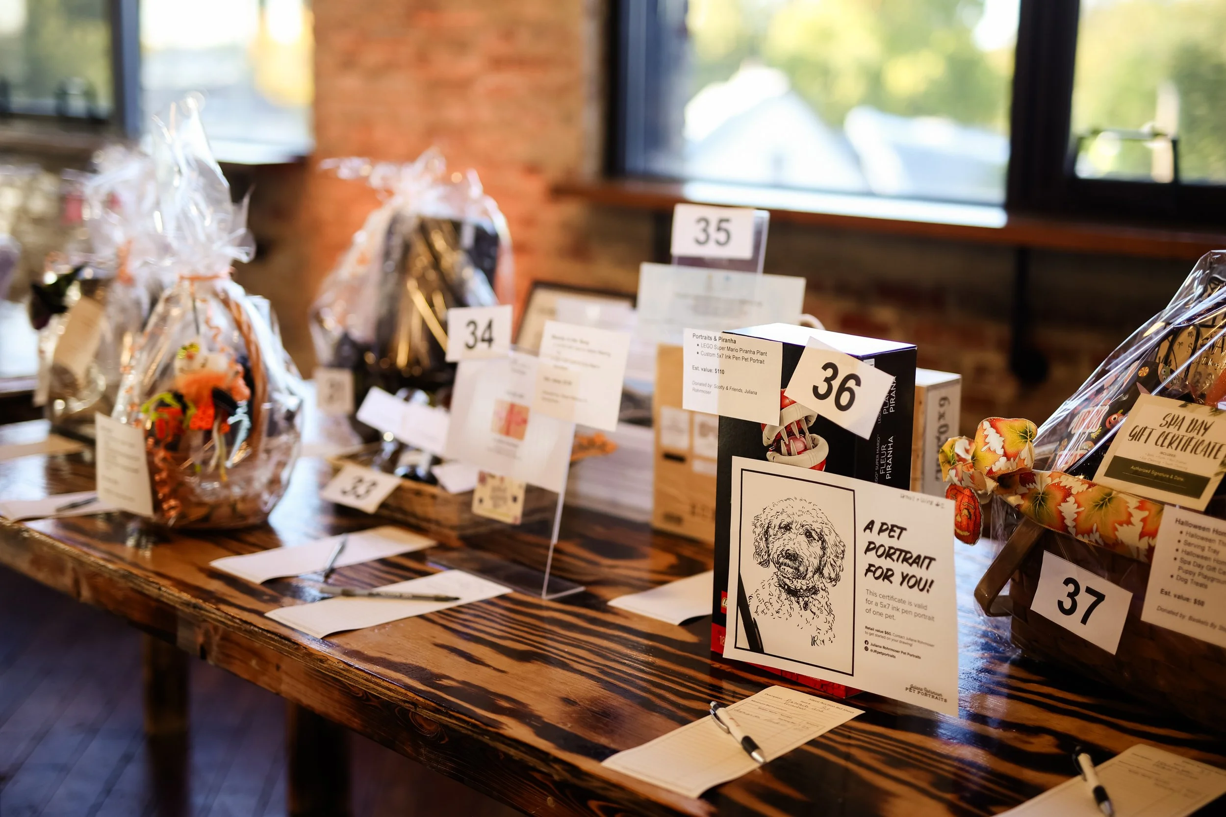 Display table with various auction items including pet portraits, gift certificates, and gift baskets, each labeled with auction lot numbers in a cozy indoor setting.