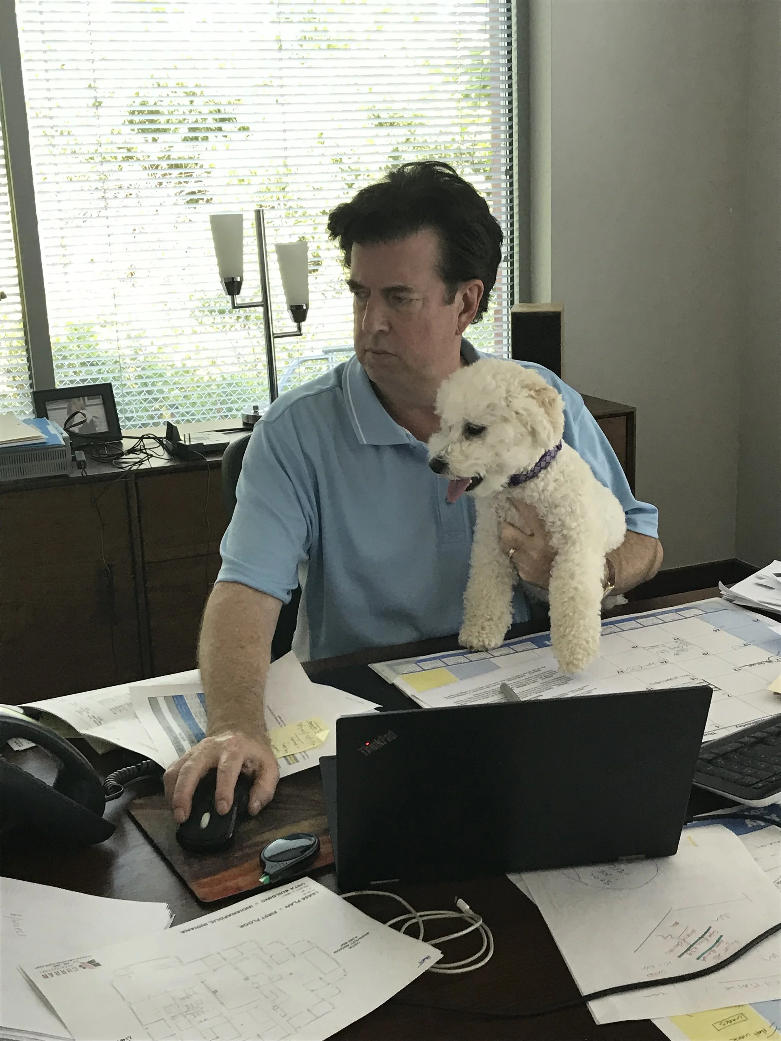 A man wearing a light blue polo shirt working at a cluttered desk with papers, a computer, a mouse, and a laptop. He is holding a small white poodle dog on his lap, while using a computer mouse.