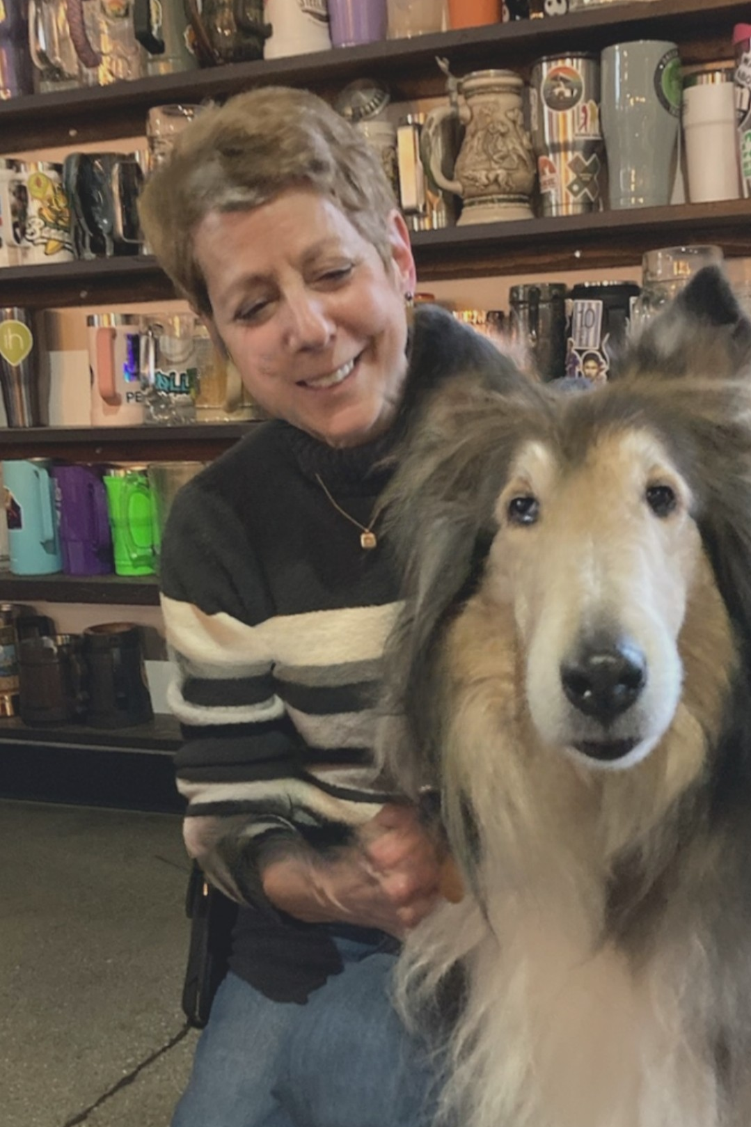 A woman smiling and petting a large dog, possibly a Rough Collie, inside a store with shelves of mugs and other items in the background.