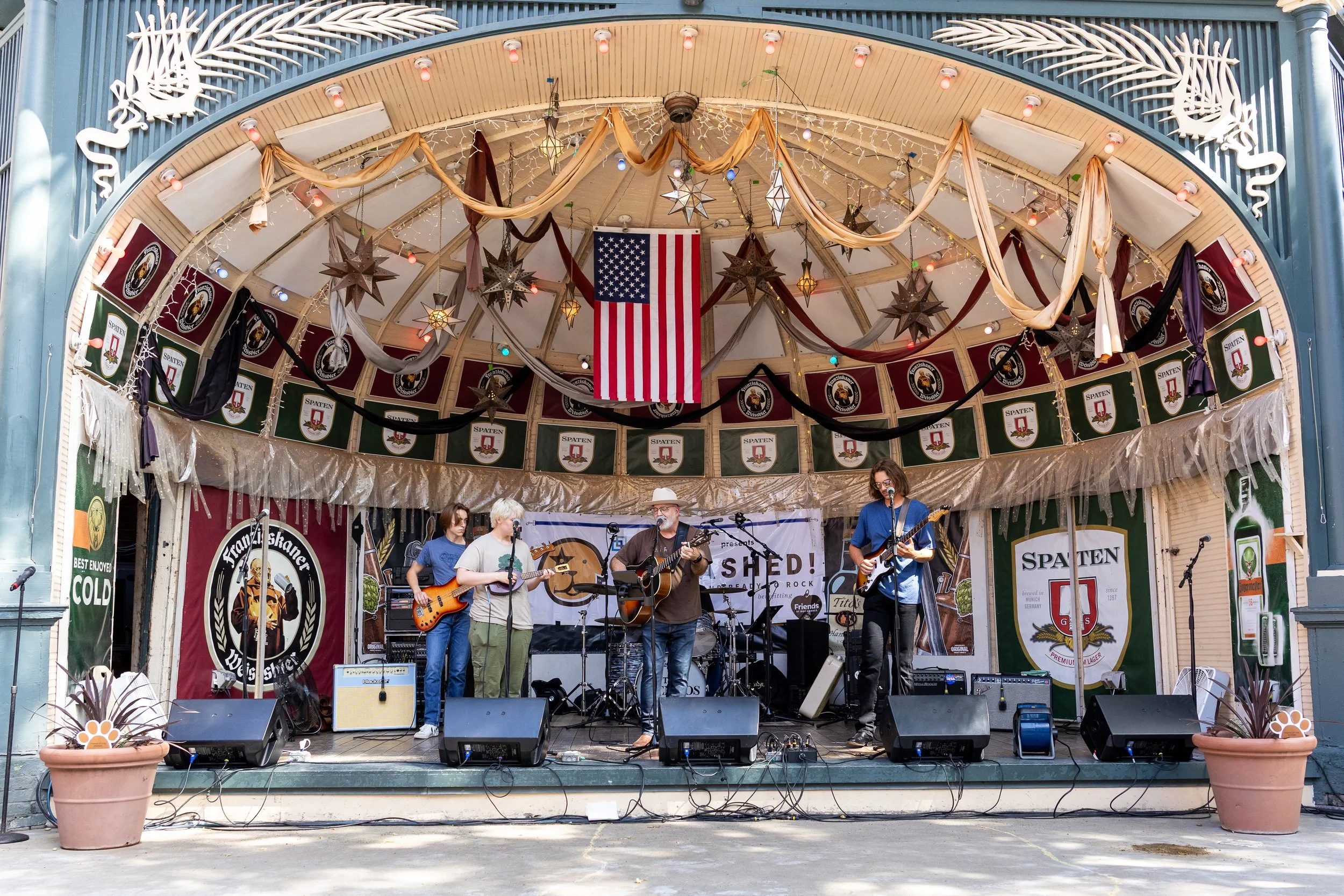 A live outdoor music performance on a decorated stage with hanging stars, draped fabric, string lights, and an American flag at the center. Musicians are playing guitars and singing into microphones, with speakers and audio equipment on the stage.
