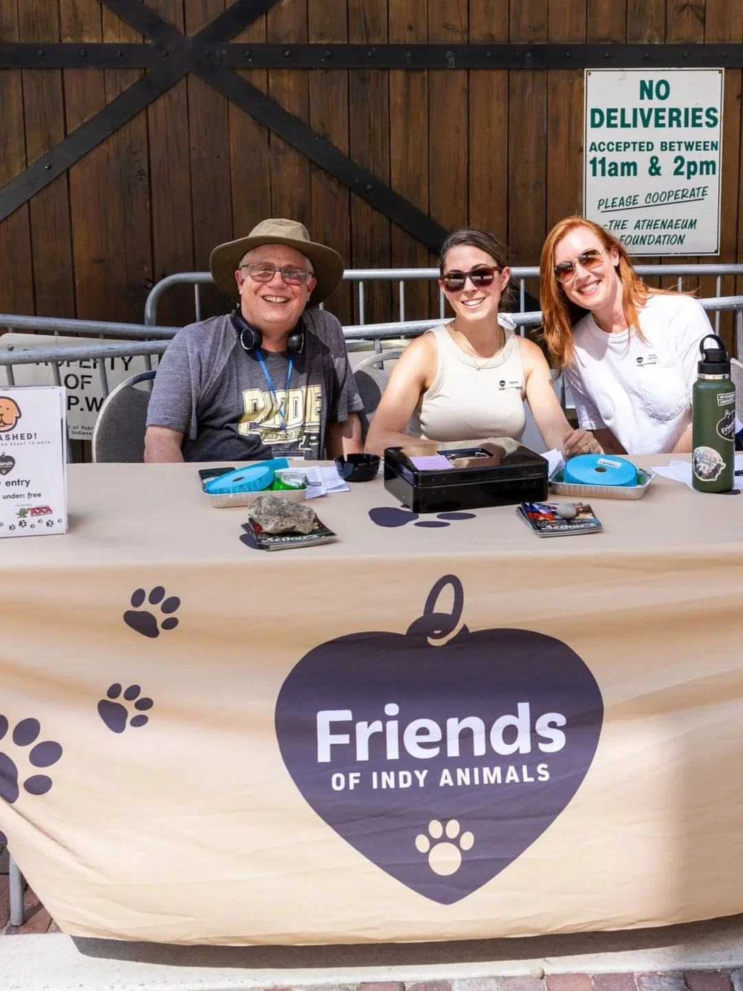 Three smiling people sit behind a table with a banner that reads "Friends of Indy Animals" at an outdoor event. The man on the left wears glasses, a hat, and a T-shirt. The two women on the right wear sunglasses and light-colored tops. The table has pet-related items and flyers.