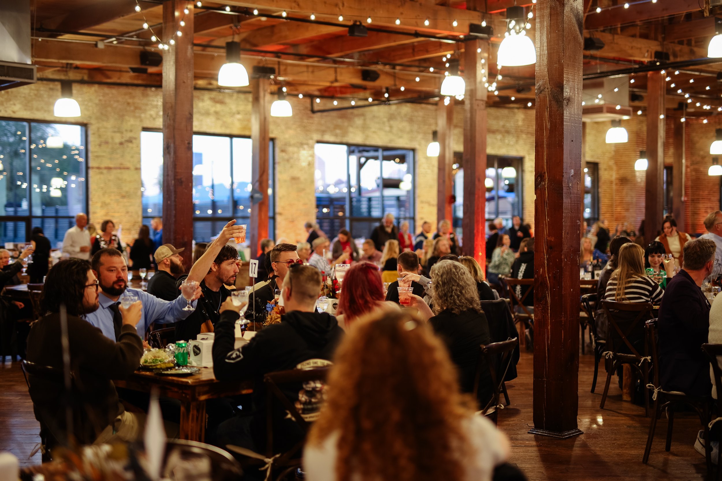 A crowded indoor event space with wooden beams and exposed brick walls, decorated with string lights and hanging pendant lights, with many people sitting at tables, some raising glasses in celebration.