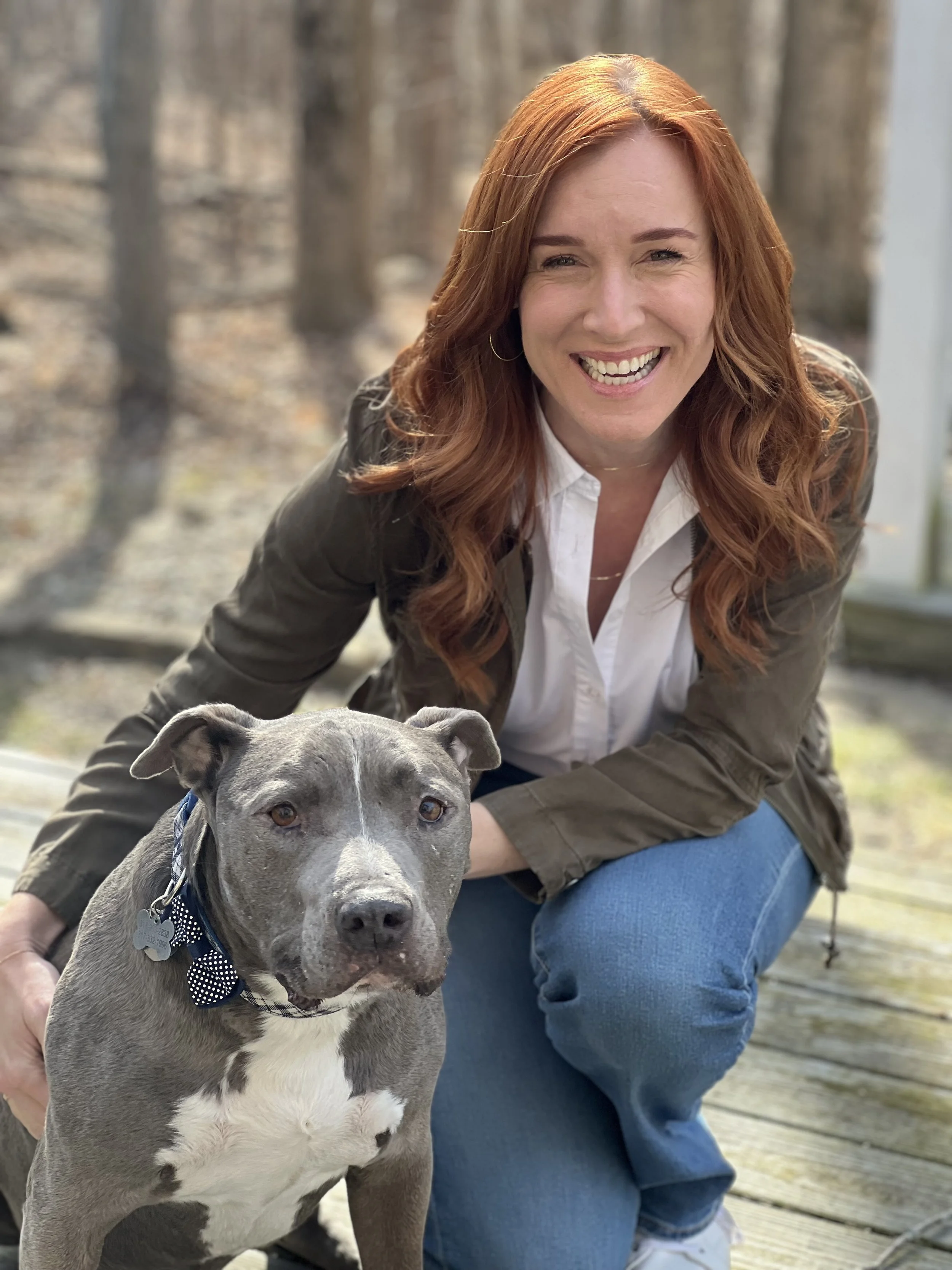 A smiling woman with red hair wearing a white shirt, brown jacket, and blue jeans kneeling on a wooden deck next to a gray and white pitbull dog with a polka-dot bow tie, outdoors with a blurred forest background.