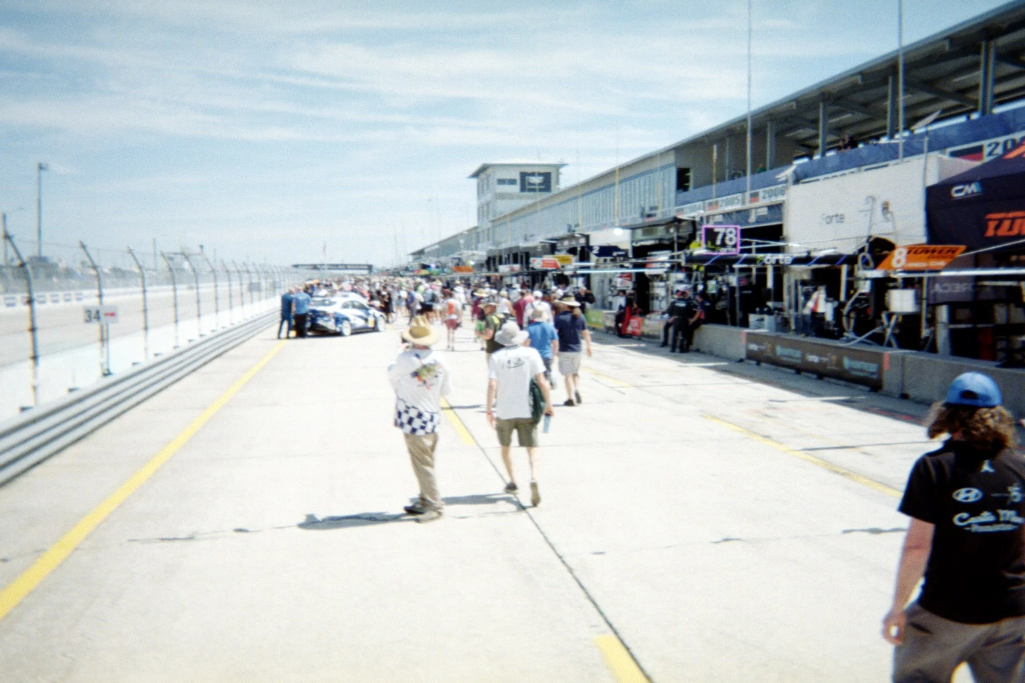 Pit row at a race track with race cars, race team garages, and people walking and working in the pit area on a sunny day.