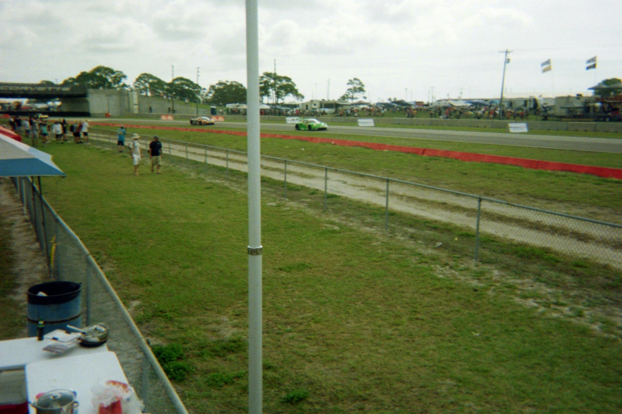 Race track with race cars speeding along the circuit, spectators watching from the side, flags flying in the background, overcast sky, and a grassy area with a chain-link fence in the foreground.