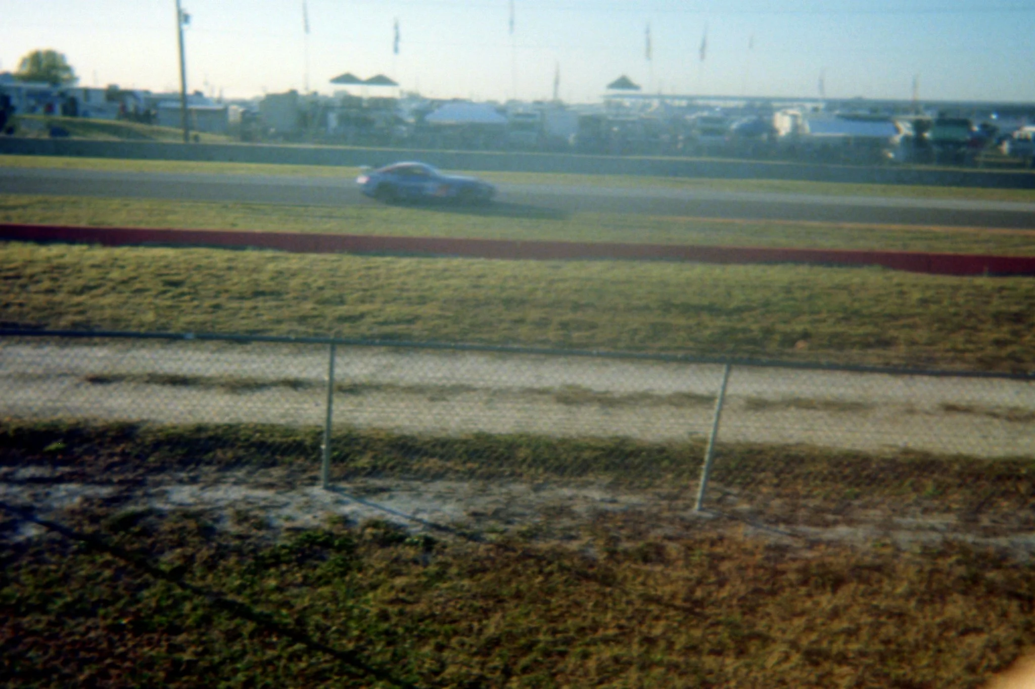 A race car moving on a track with grass and a chain-link fence in the foreground, and an area with parked trailers, tents, and poles in the background.