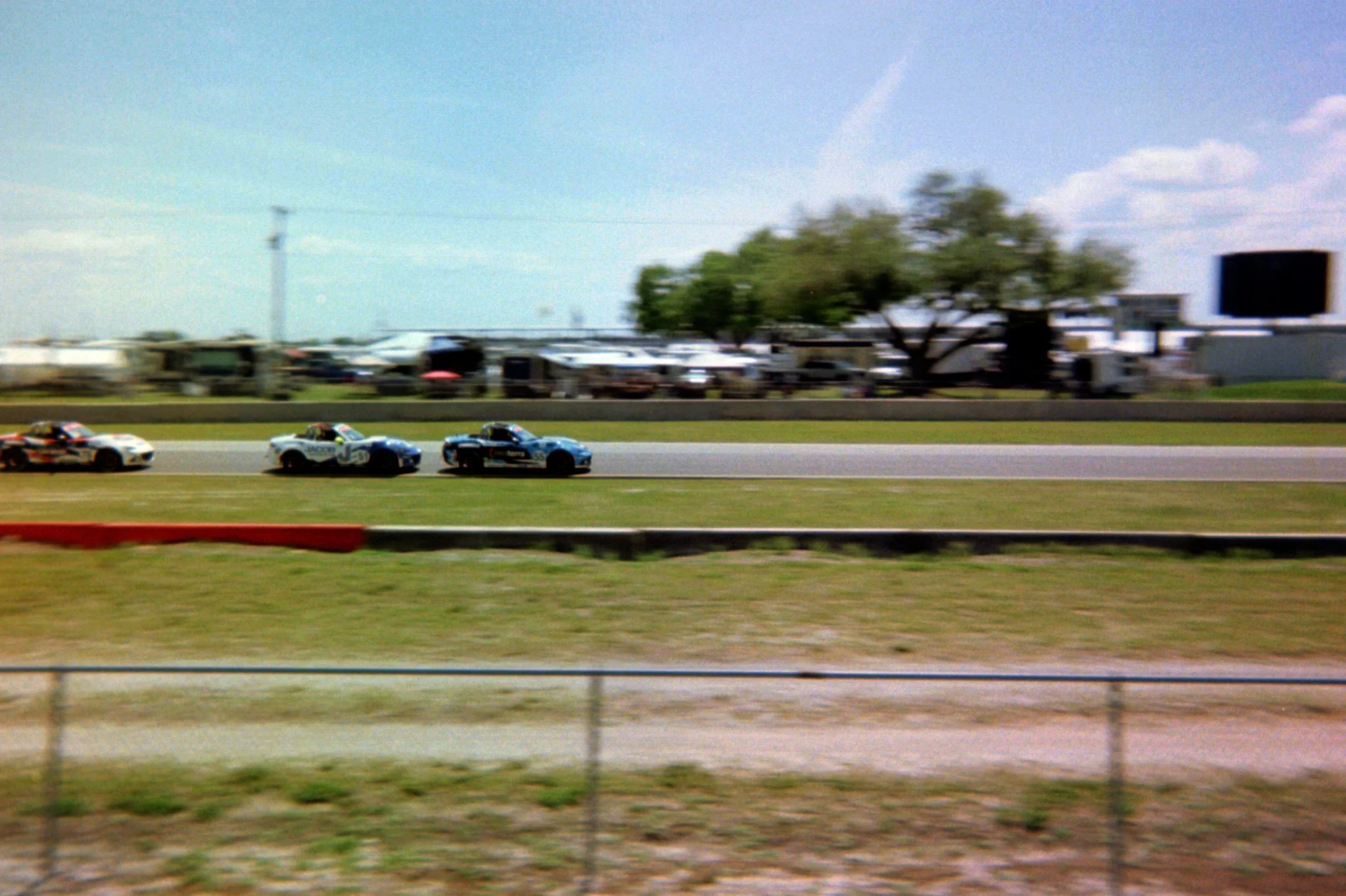 Race cars racing on a track during daytime with blurred background of trailers and trees.