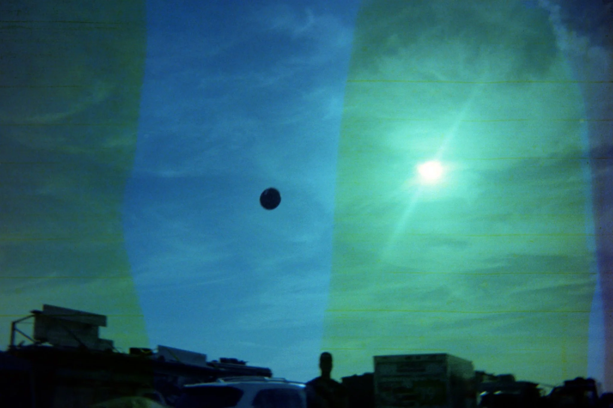 A surreal photo showing a bright sun, a small dark object (possibly a moon or planet), and a silhouette of a person standing on the ground with objects and cars around, under a sky with unusual green and blue hues.