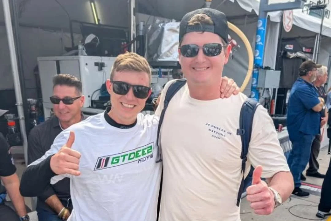 Three men at an outdoor event, smiling and giving thumbs-up signs. All are wearing sunglasses, and two have backpacks. Background shows tents, equipment, and other people.