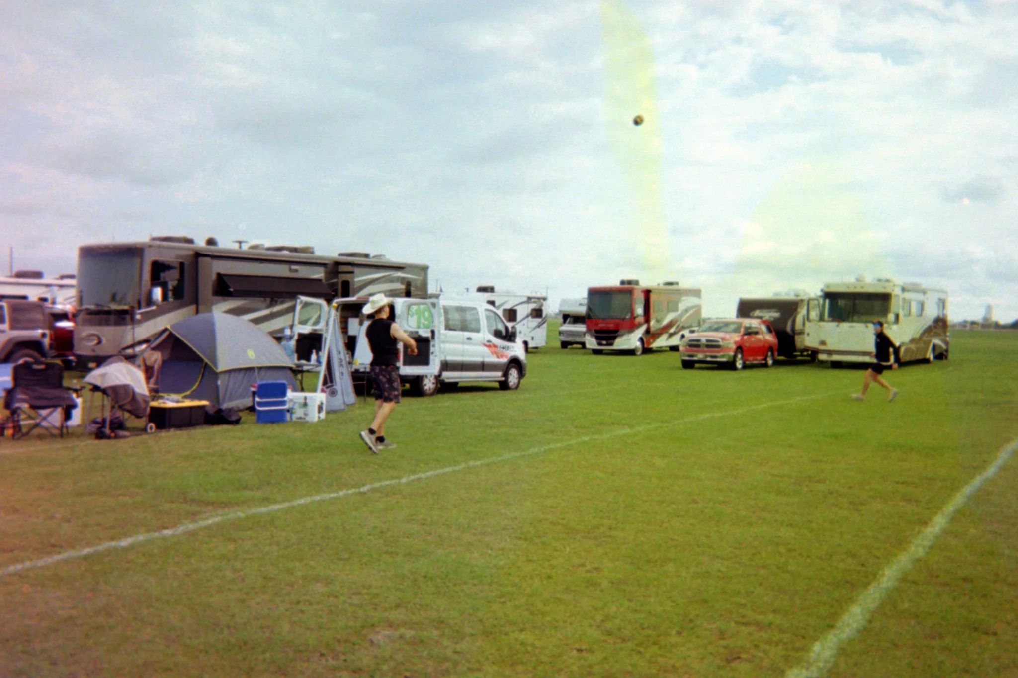 A grassy field with RVs, camping tents, and cars parked in a row. Two people are walking, one near the tents and another near the RVs. The sky is partly cloudy.