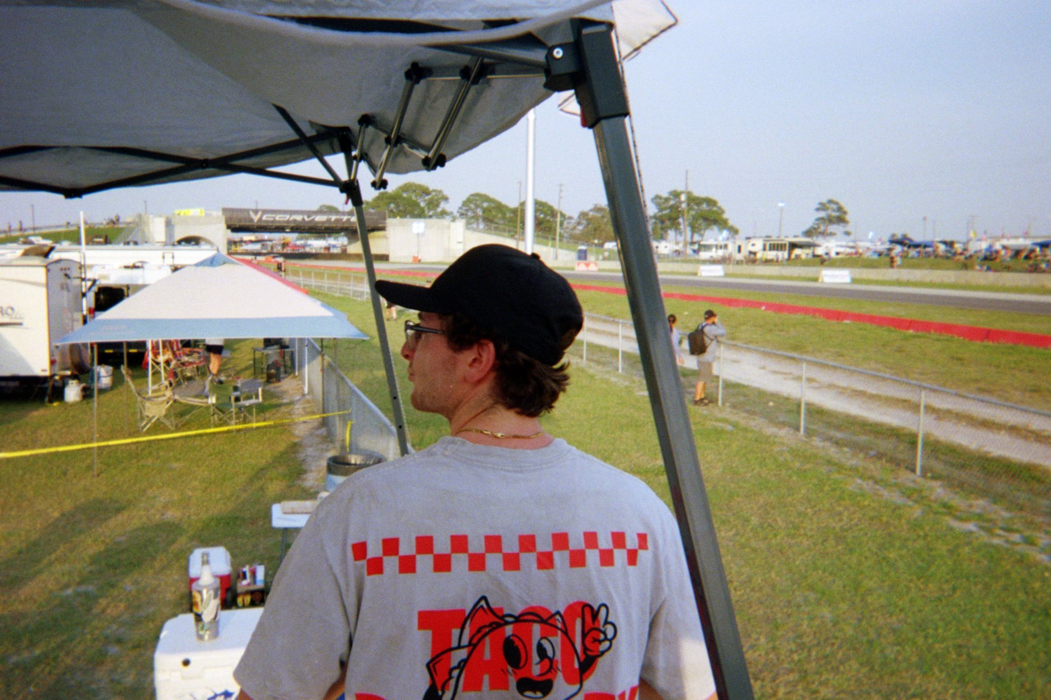 A man wearing a black cap, glasses, and a white T-shirt with a red checkered pattern and a cartoon mascot on the back, standing under a canopy at an outdoor event near a race track during daytime.