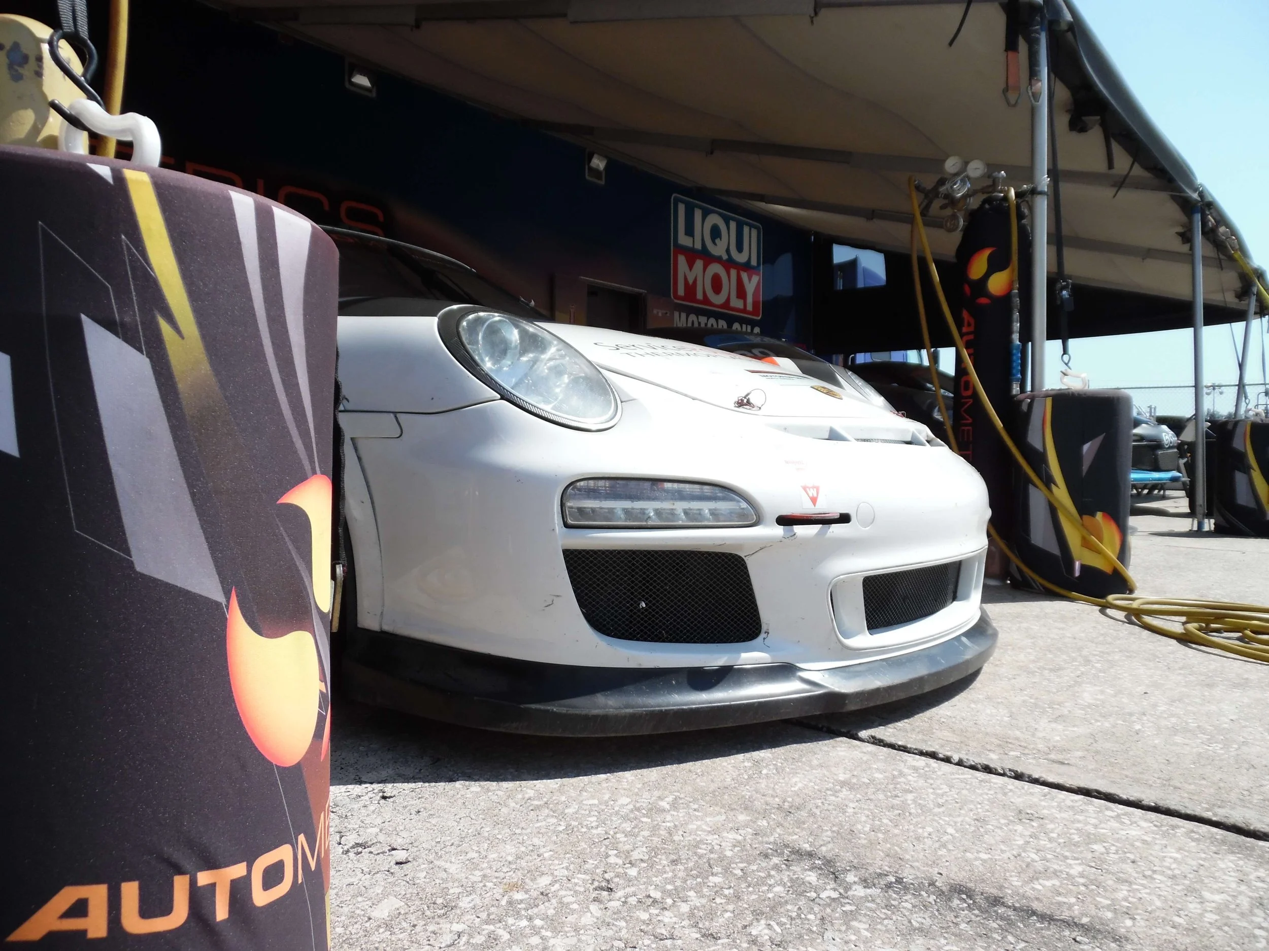 White race car in a pit garage with a black front splitter, headlight, and hood vents, surrounded by yellow air hoses, with Liqui Moly banners in the background.