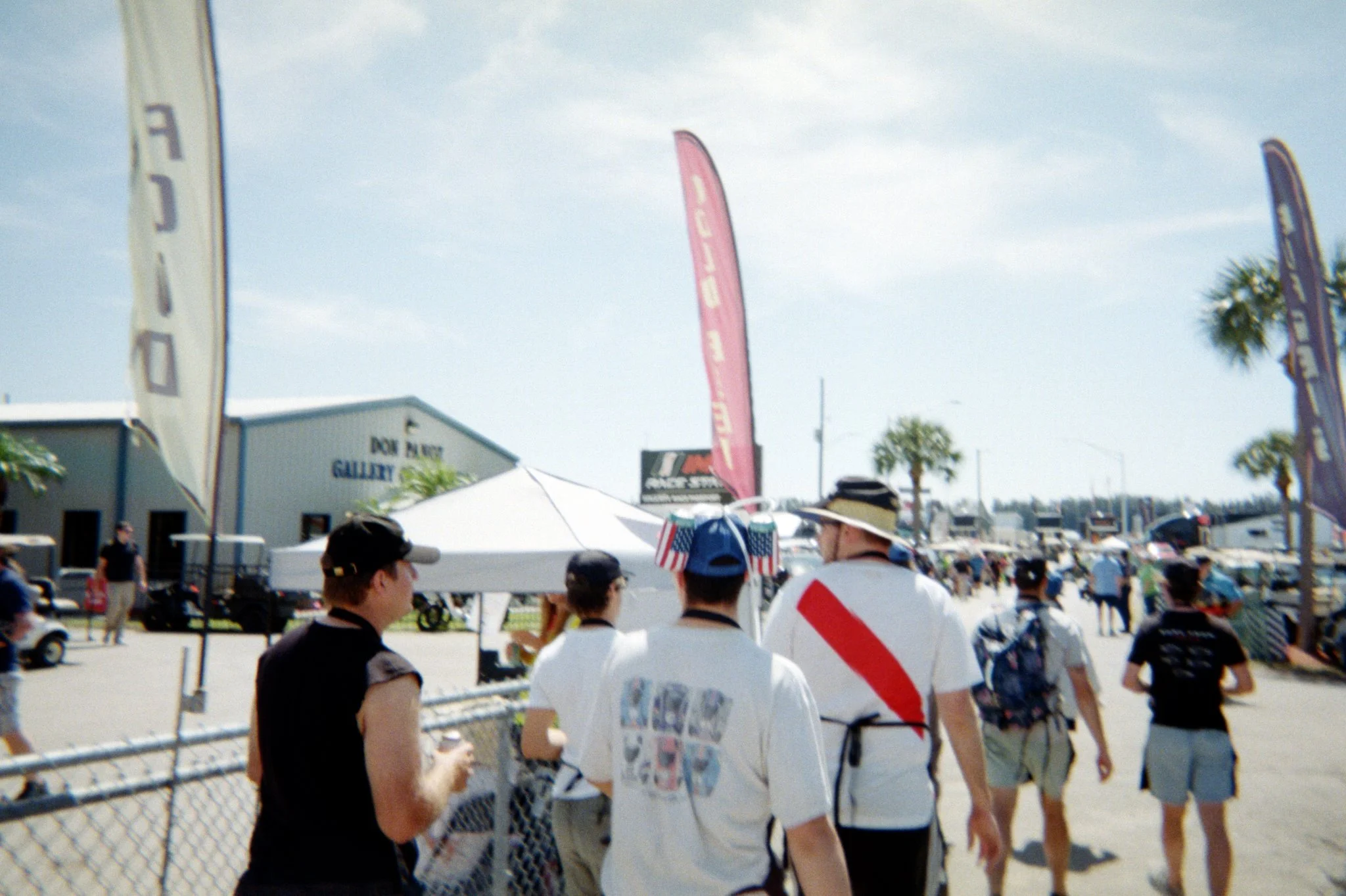 People walking at an outdoor event with flags, tents, and a building in the background, under a partly cloudy sky.