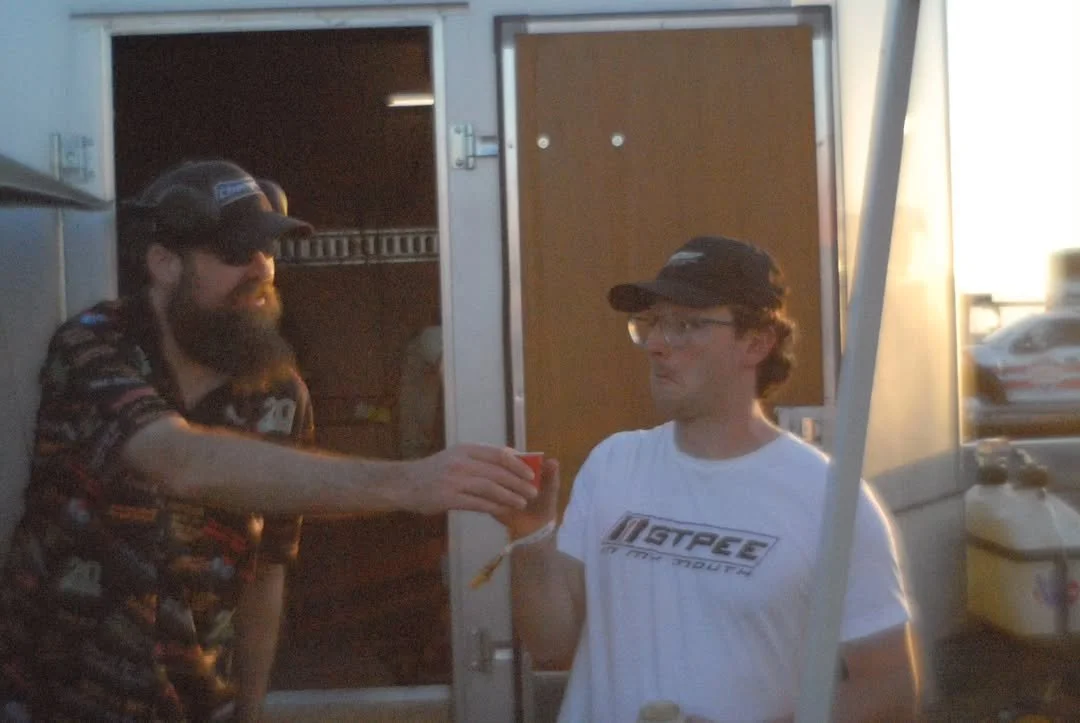 Two men standing outside near a food truck; one man is giving a drink to the other.