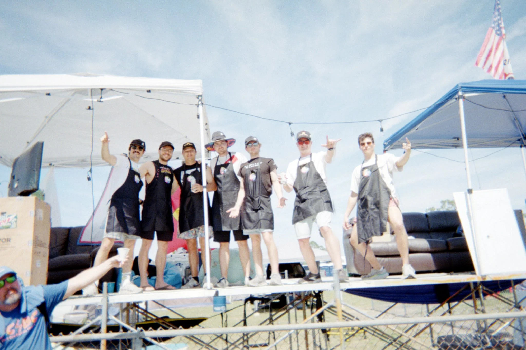 Group of seven people on a stage outdoors under blue skies, some wearing aprons, some holding drinks, making hand gestures, and posing for the photo. Two tents and American flags are visible in the background.