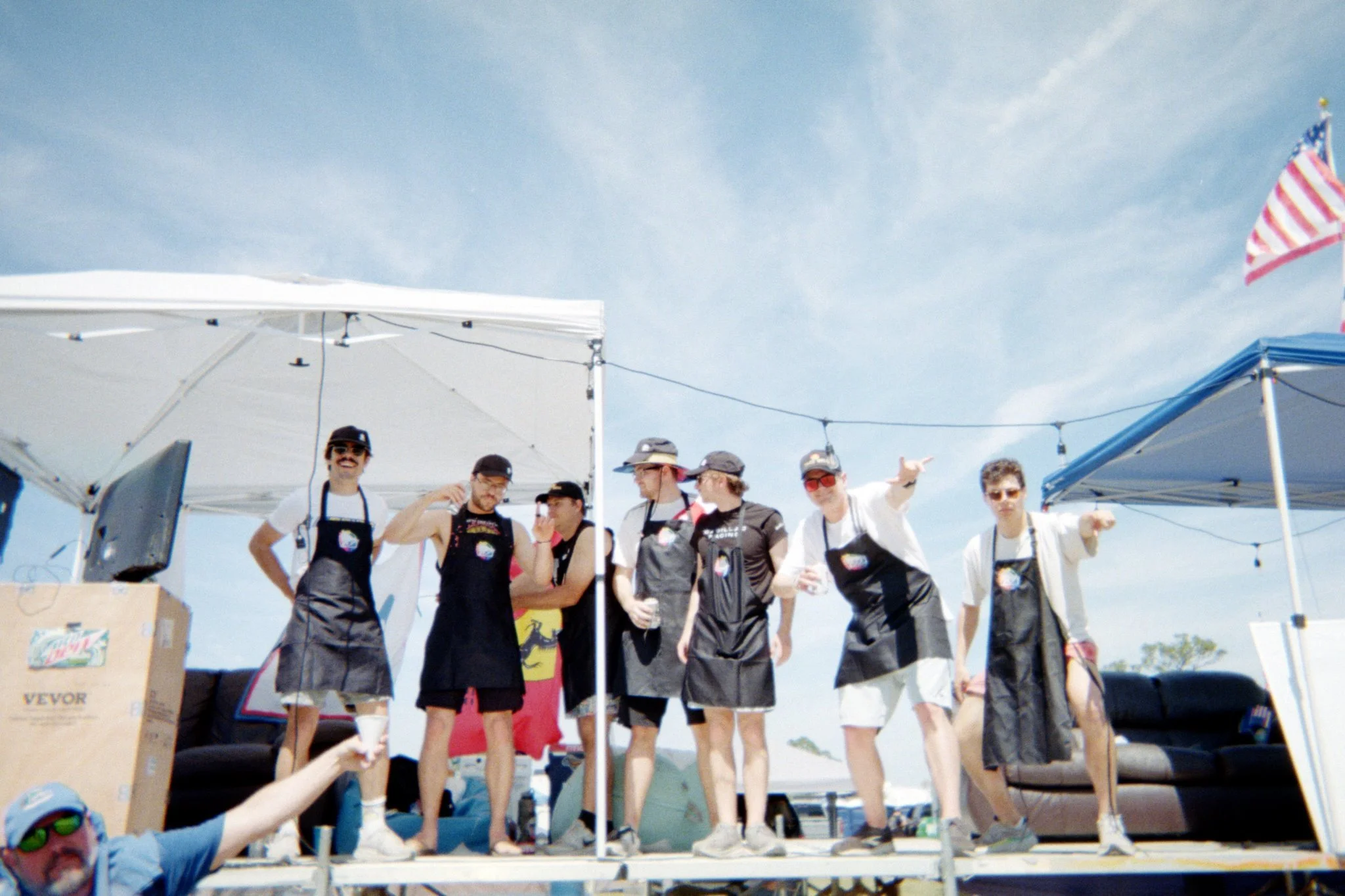 Group of people at an outdoor event, some wearing aprons, standing on a stage under tents with a blue sky in the background.