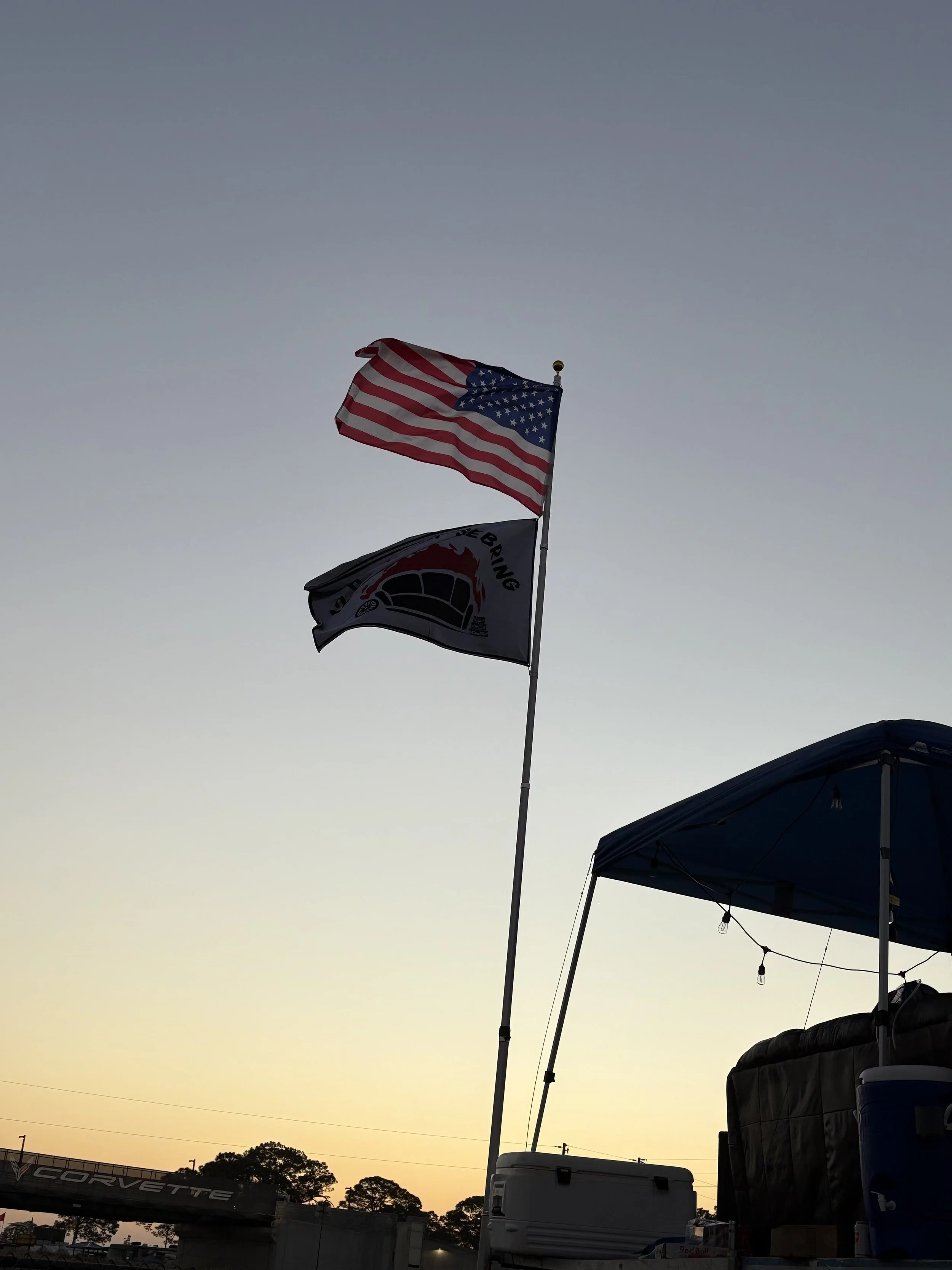 American flag and a second flag flying at sunset near a tent and cooler.
