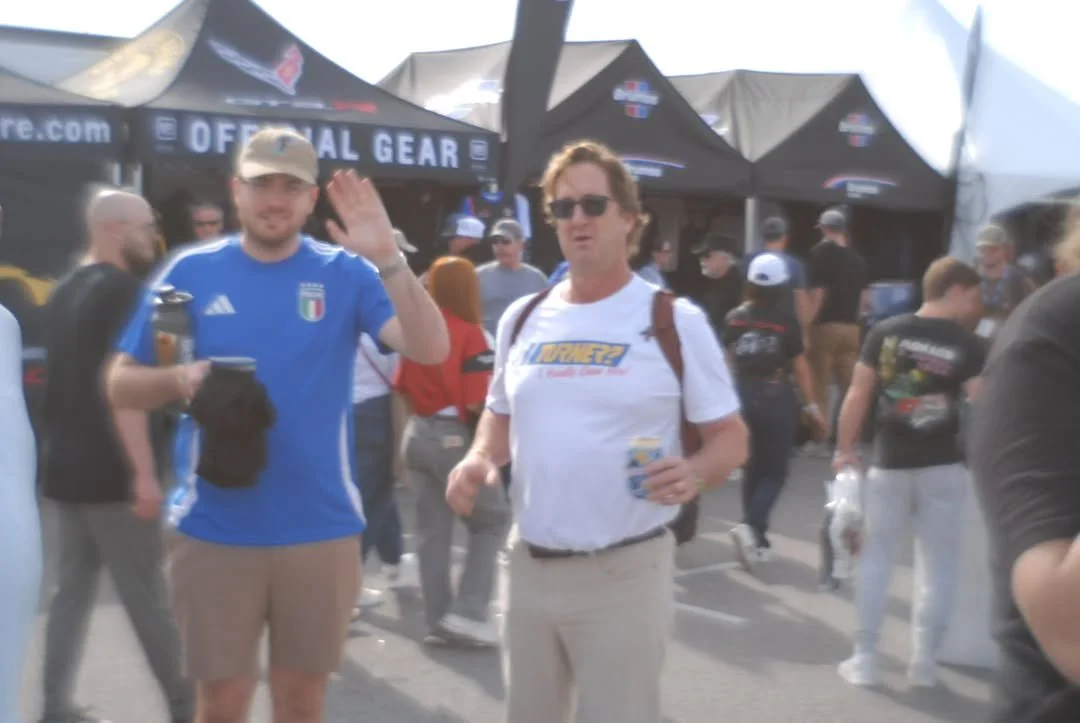 Two men waving at the camera at an outdoor event with tents and several people in the background