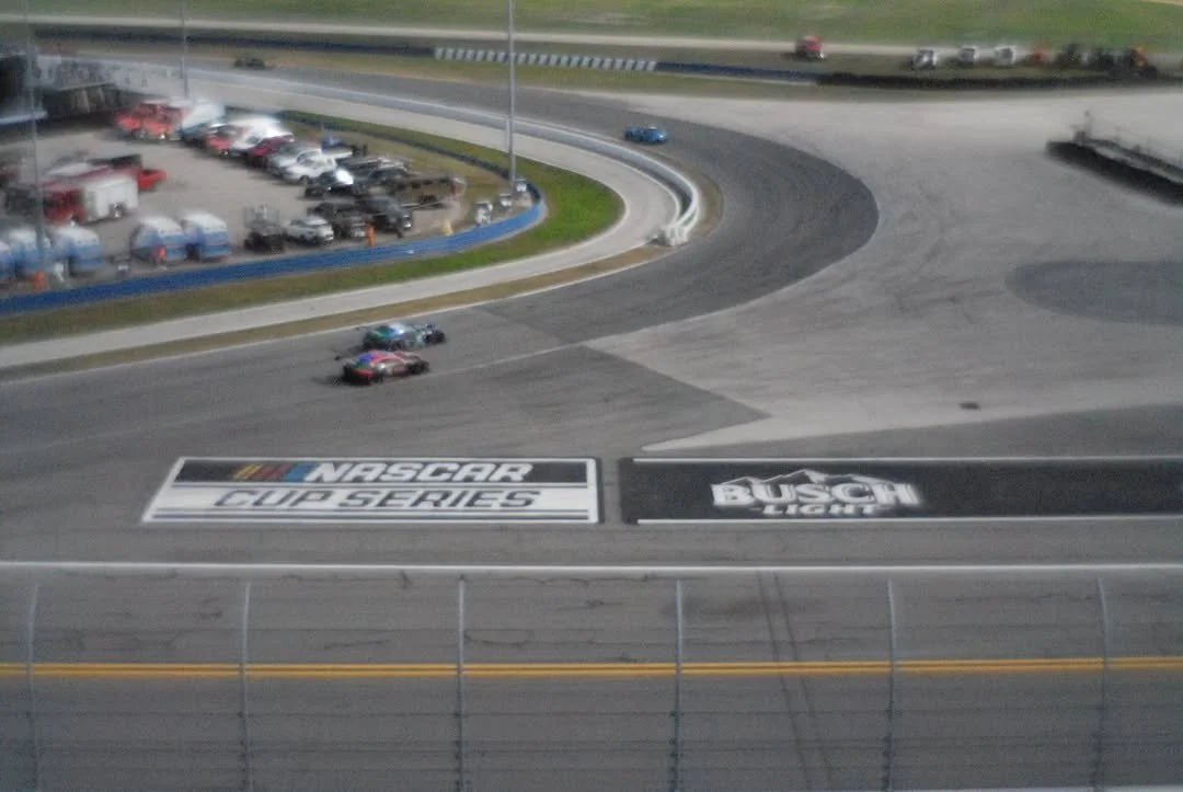 Two race cars on a Nascar track during a race, with NASCAR Cup Series and Busch Light signs painted on the track. There are parked cars and trucks along the outside of the track.
