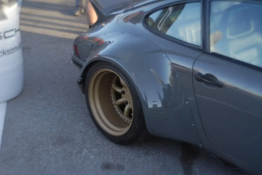 Close-up of a silver sports car with a wide wheel, parked in a lot next to a white barrel.